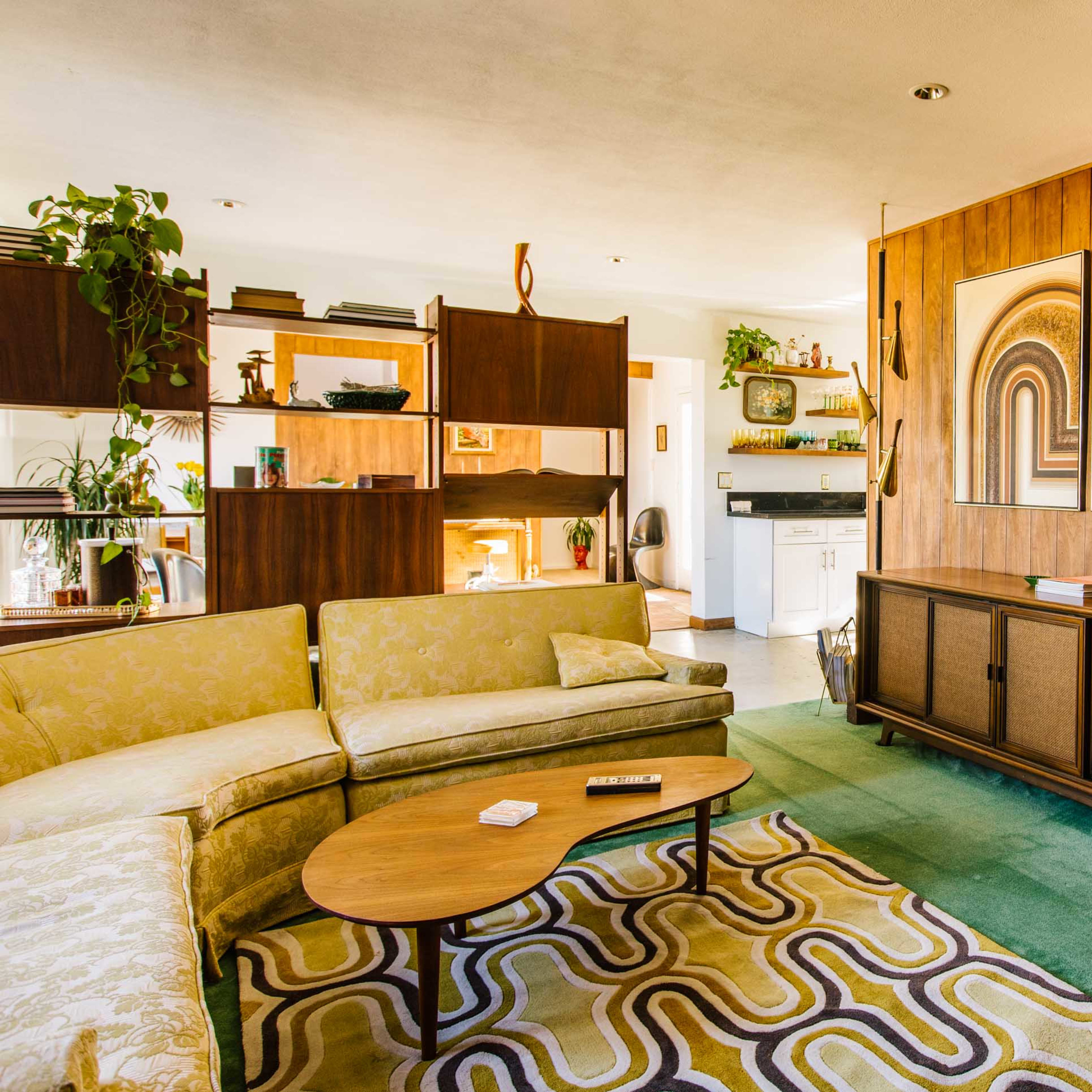 A mid-century modern living room featuring a green carpet, a patterned area rug, a sectional sofa, and framed artwork on the wooden-paneled wall.
