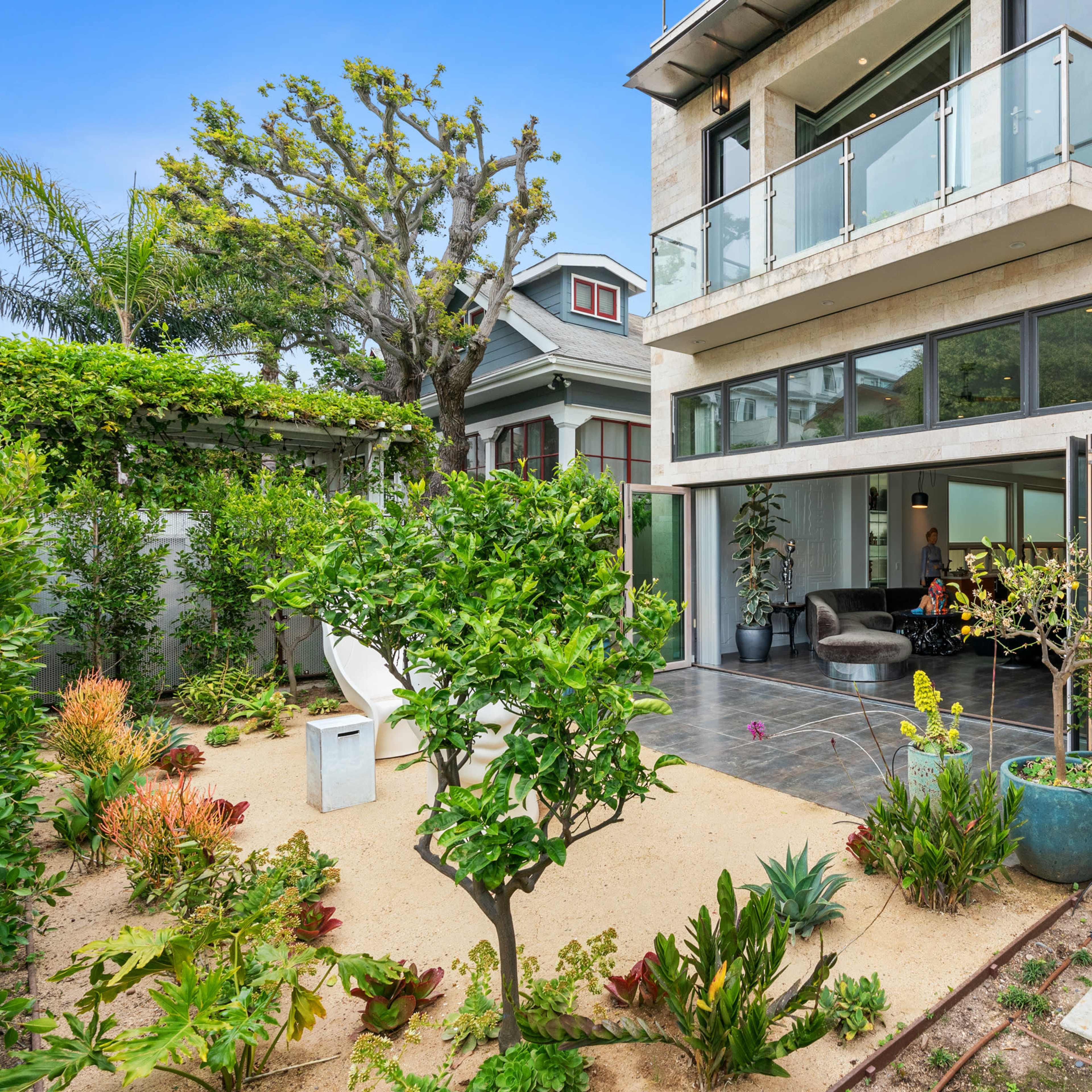 A modern two-story house with large windows, surrounded by a landscaped garden featuring small trees and plants.