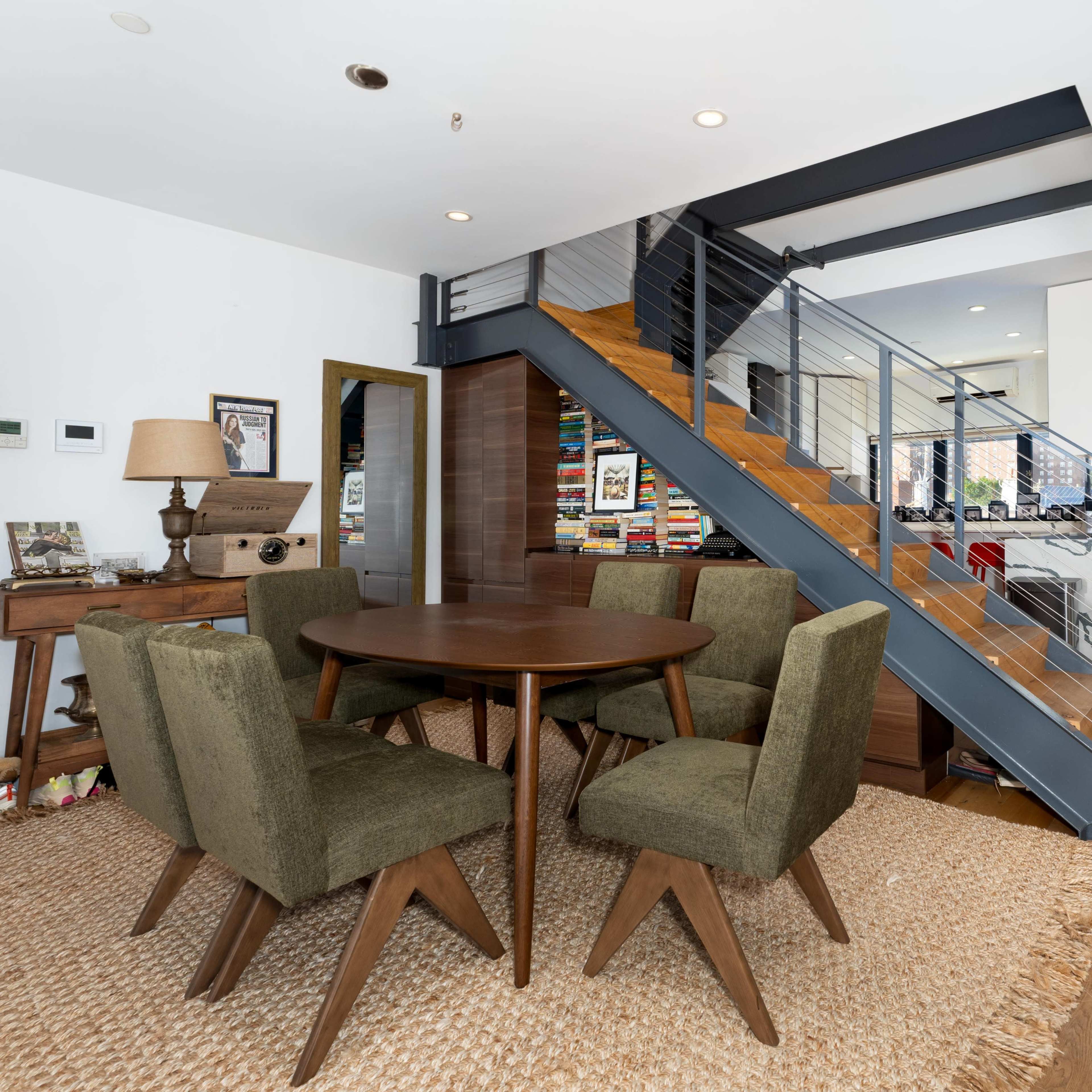 A dining area features a round wooden table surrounded by four green upholstered chairs, positioned near a modern staircase with wooden steps and a metal railing.