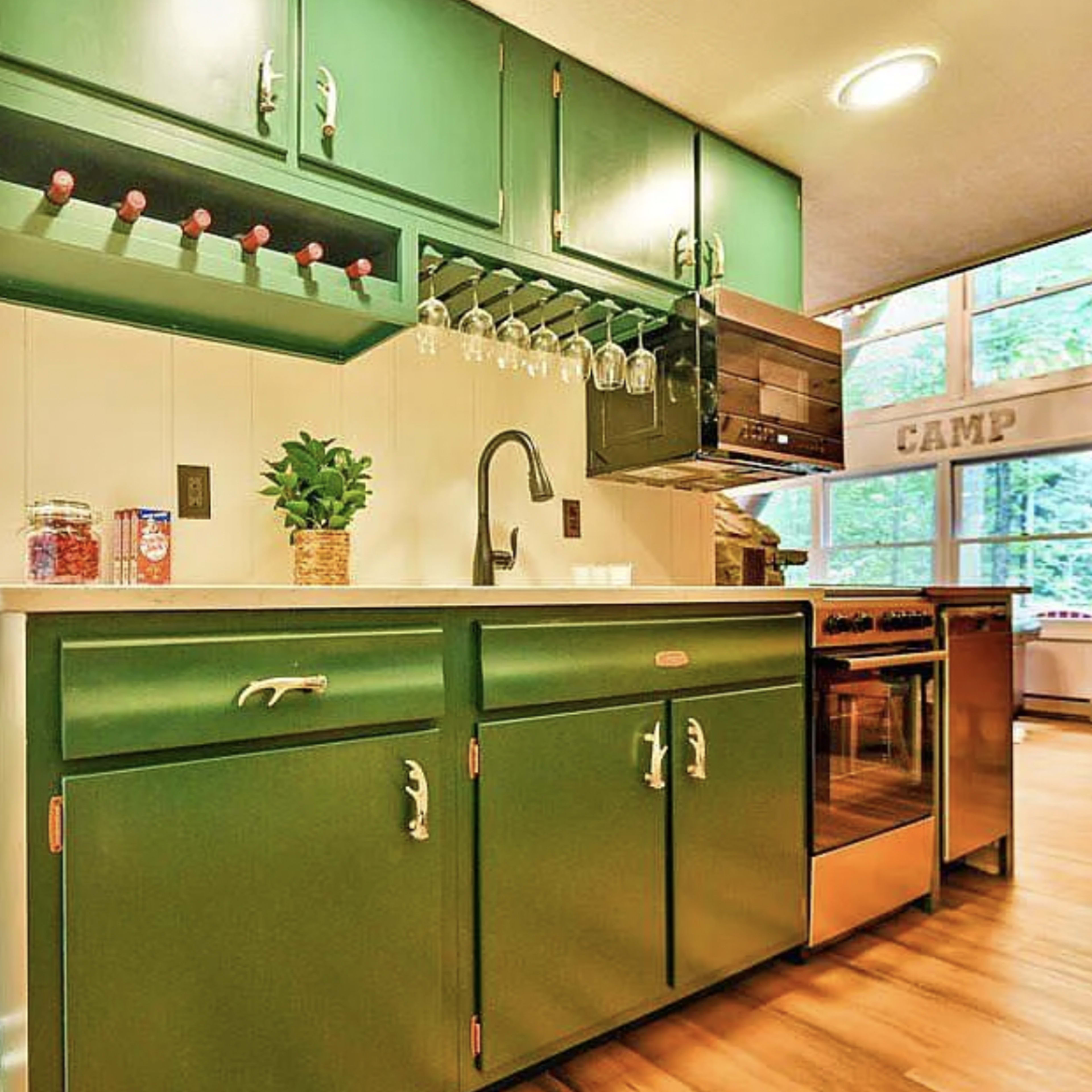 A compact kitchen with green cabinetry, a sink, and stainless steel appliances, adjacent to a bedroom area with wood-paneled walls.