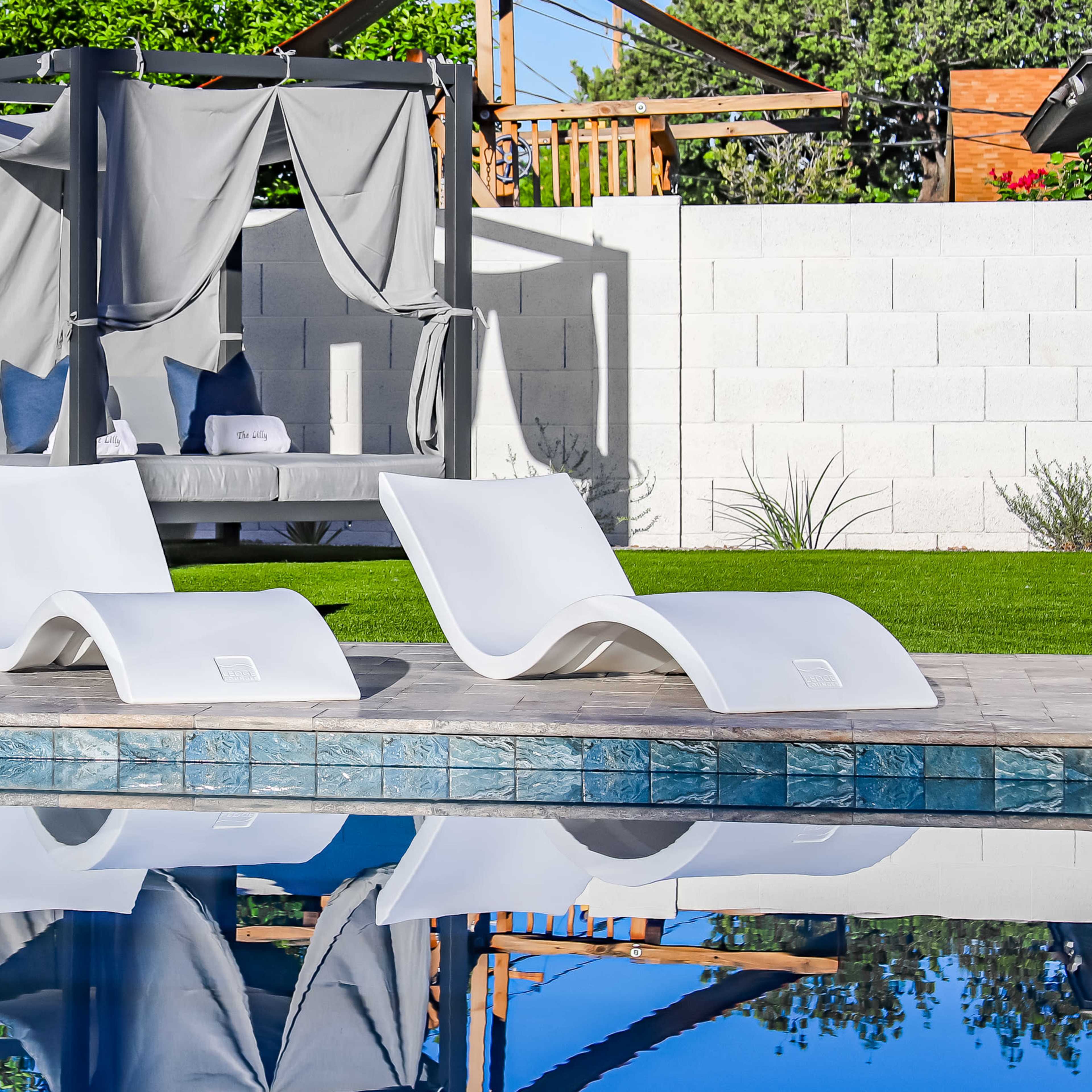 Two white lounge chairs beside a swimming pool, with a canopy-covered seating area in the background and green grass surrounding the setting.