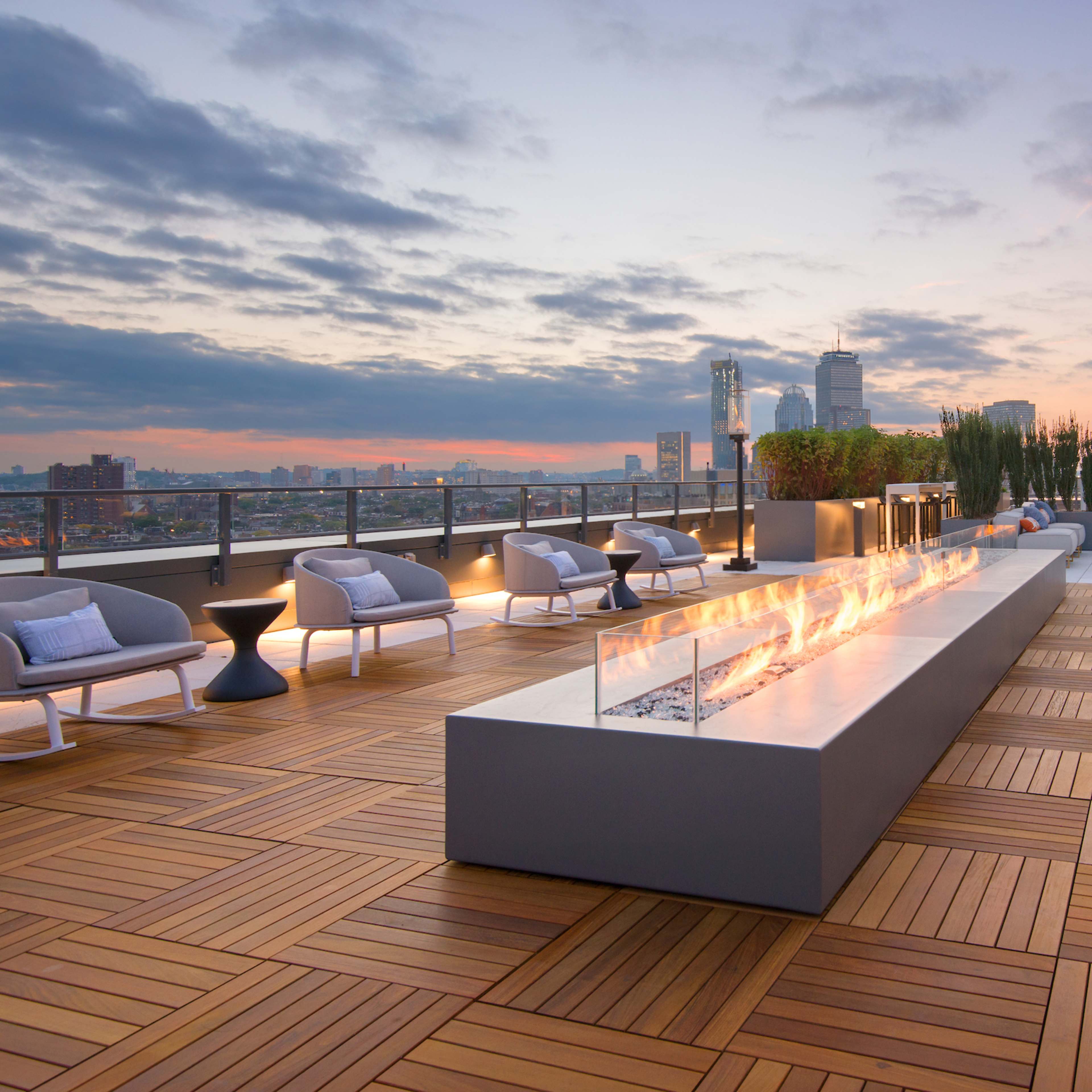 A modern living room features a sectional sofa and a bar-style seating area, with large windows offering a cityscape view and a linear fireplace along one wall.