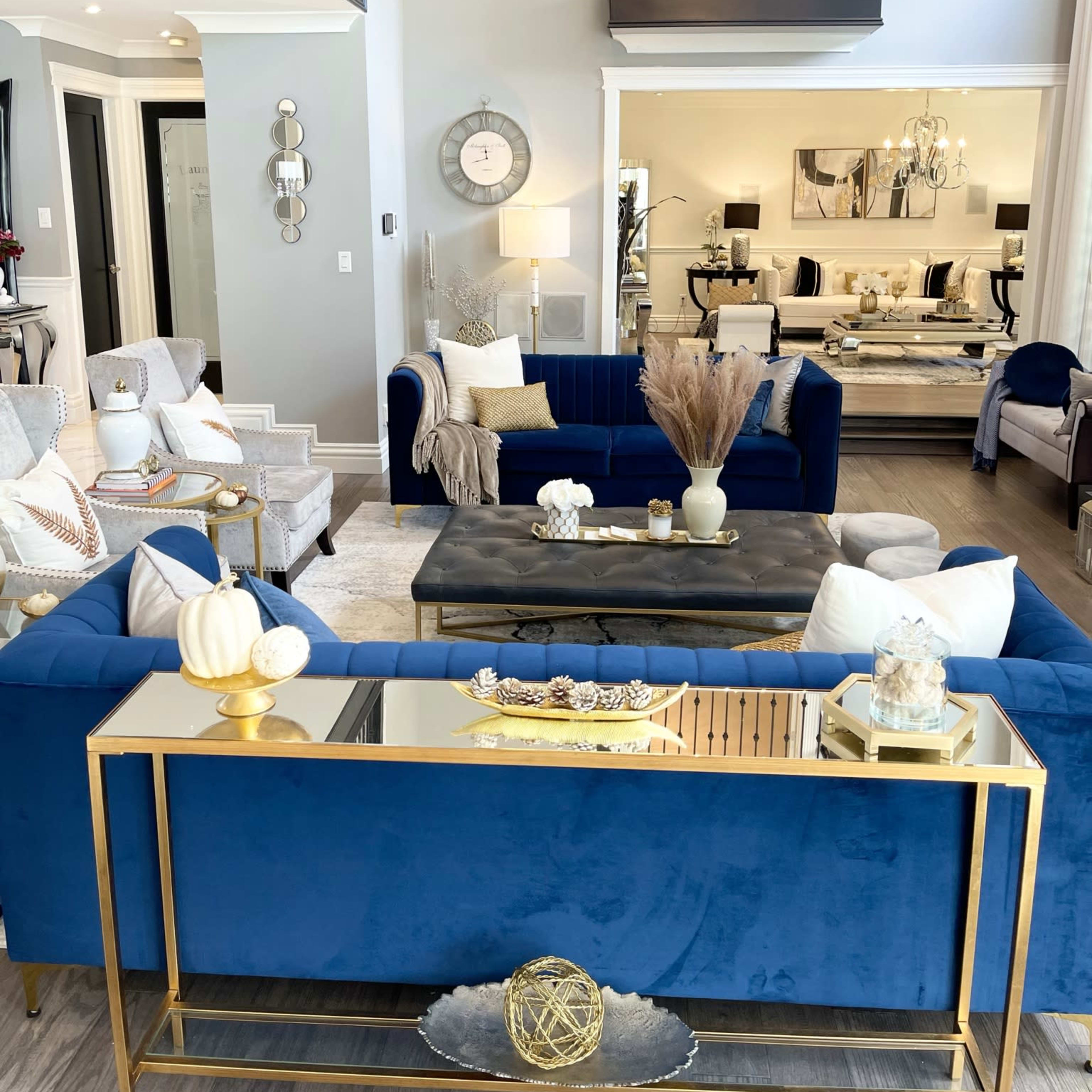 A modern living room with blue velvet sofas, a glass-top coffee table, and decorative elements like vases and a mirror, set in a spacious area with high ceilings and a balcony above.