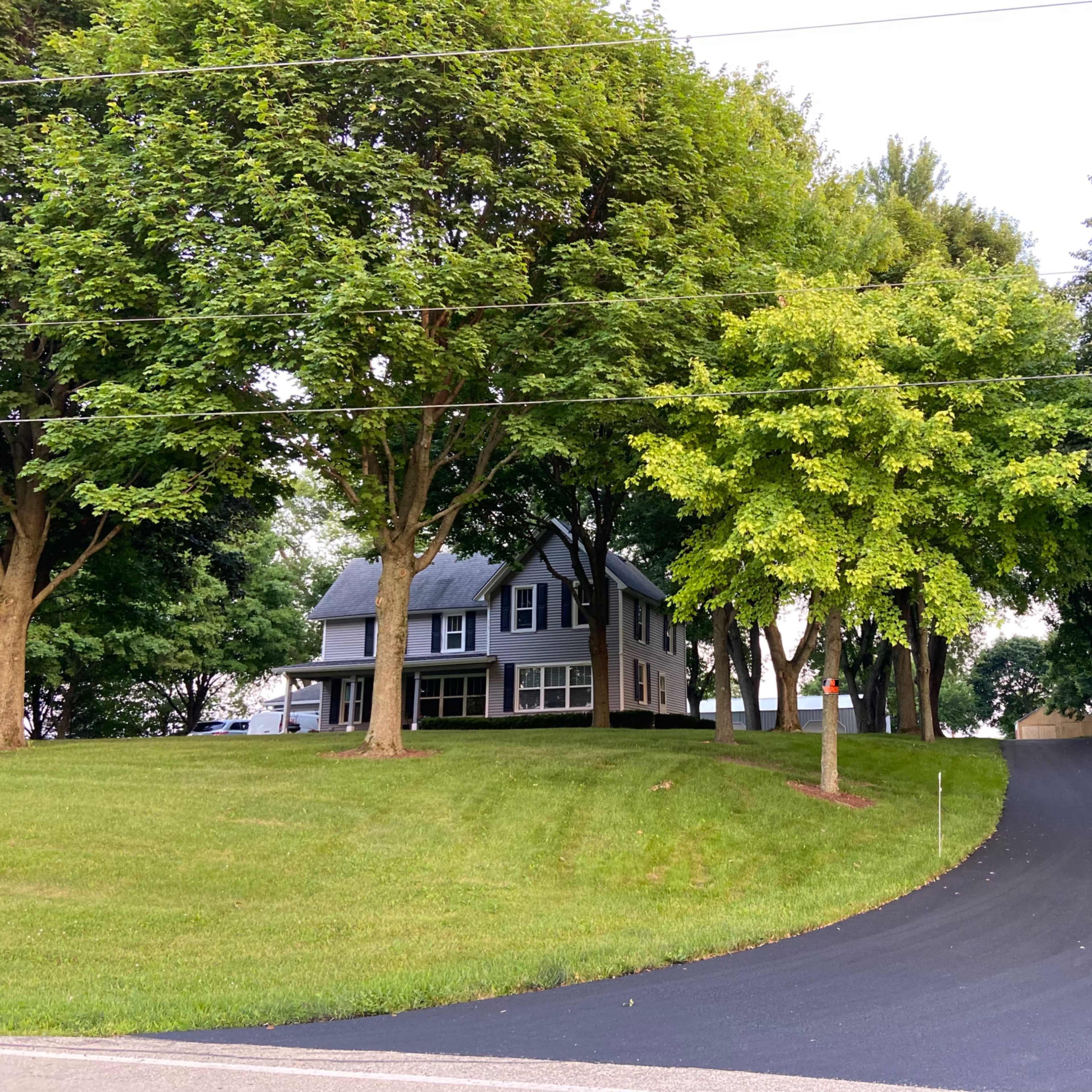A grey house is situated on a grassy lot, surrounded by several trees, with a newly paved driveway leading to it.