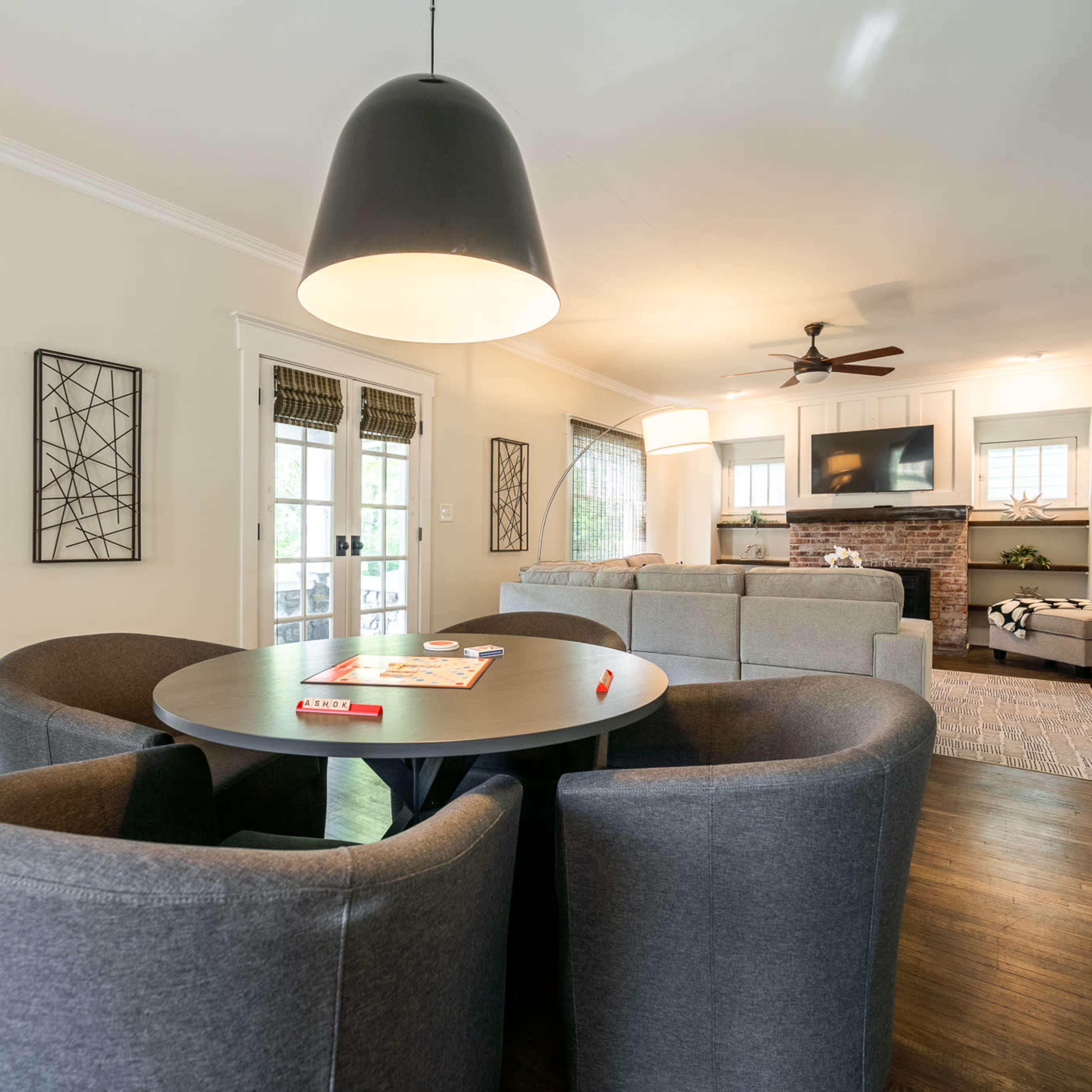 A modern living room features a circular table with four gray chairs, surrounded by natural light from large windows and a spacious couch in the background.