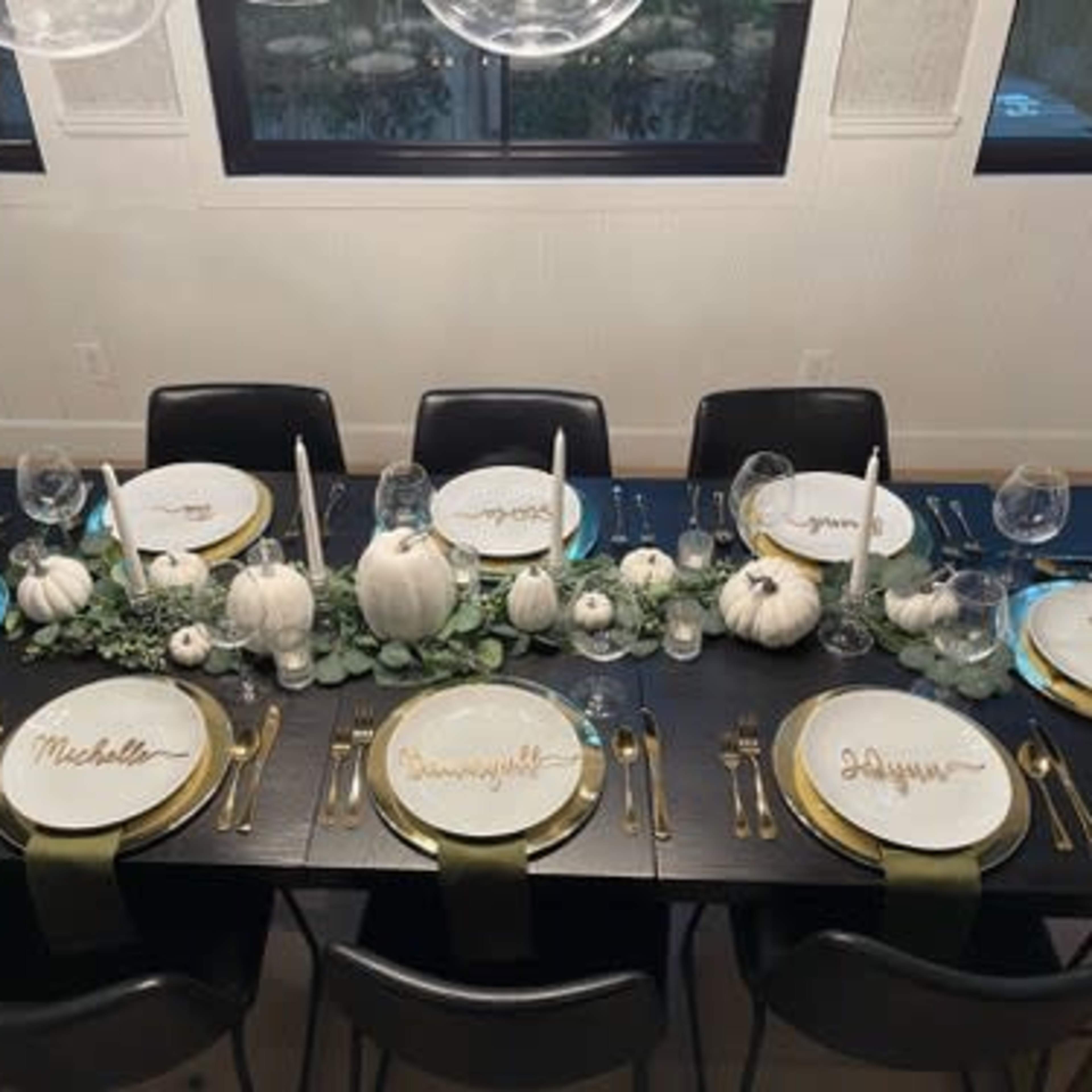 A dining table is elegantly set with white plates featuring gold names, surrounded by small white pumpkins and greenery, illuminated by overhead lights.