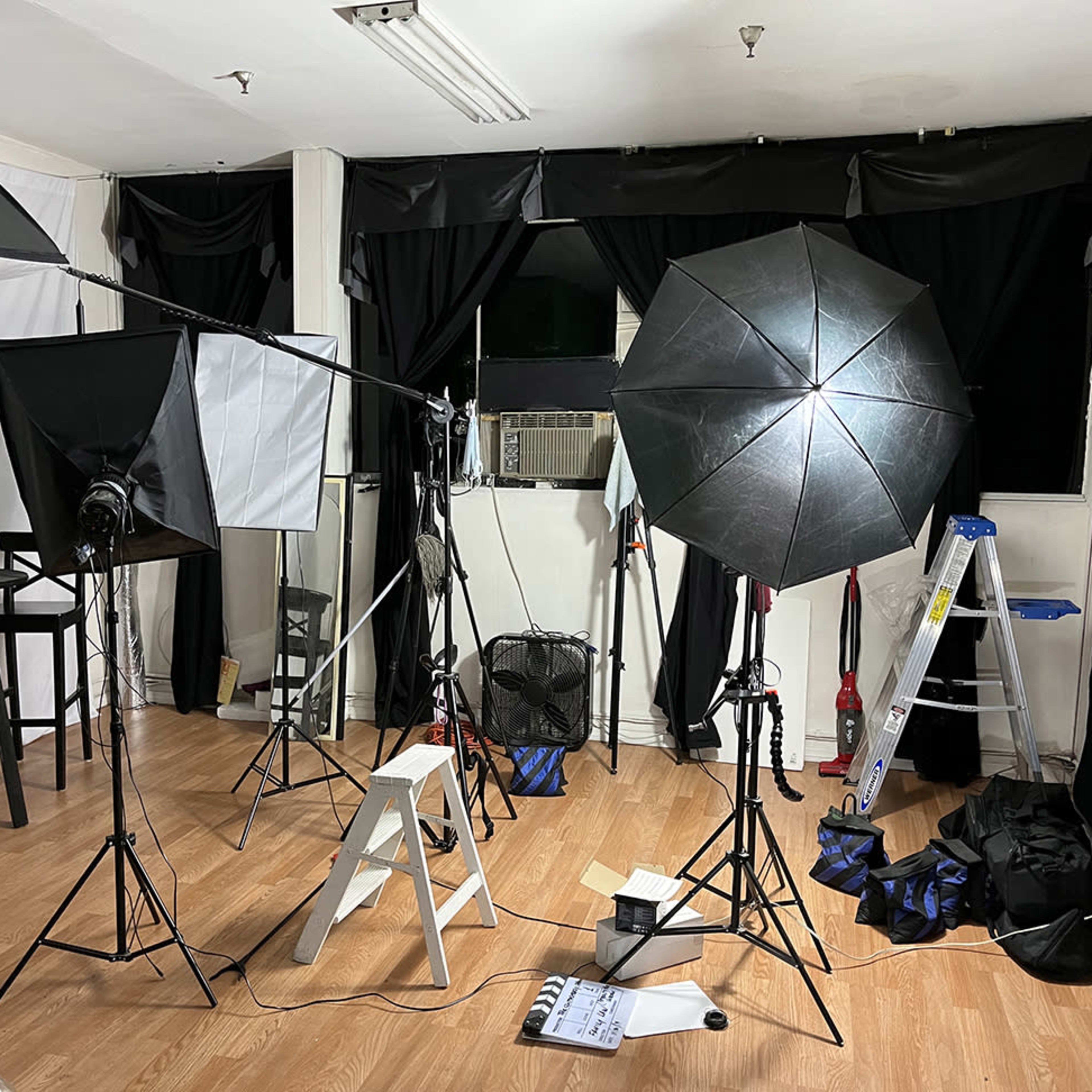 A photography studio with various lighting equipment, including softboxes and umbrellas, set up among black curtains and a wooden floor.
