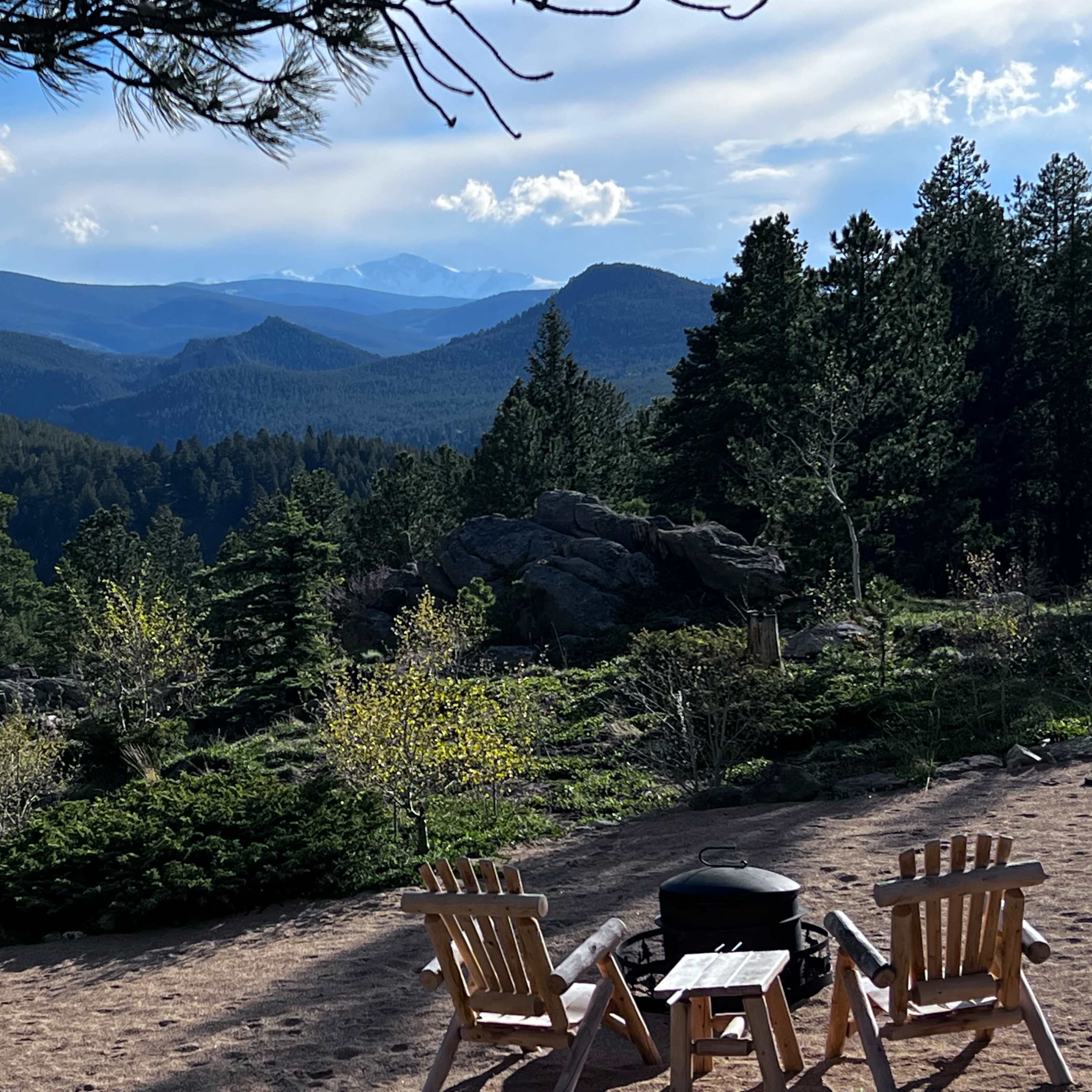 Two wooden chairs face a vast mountainous landscape dotted with trees and distant peaks under a partly cloudy sky.
