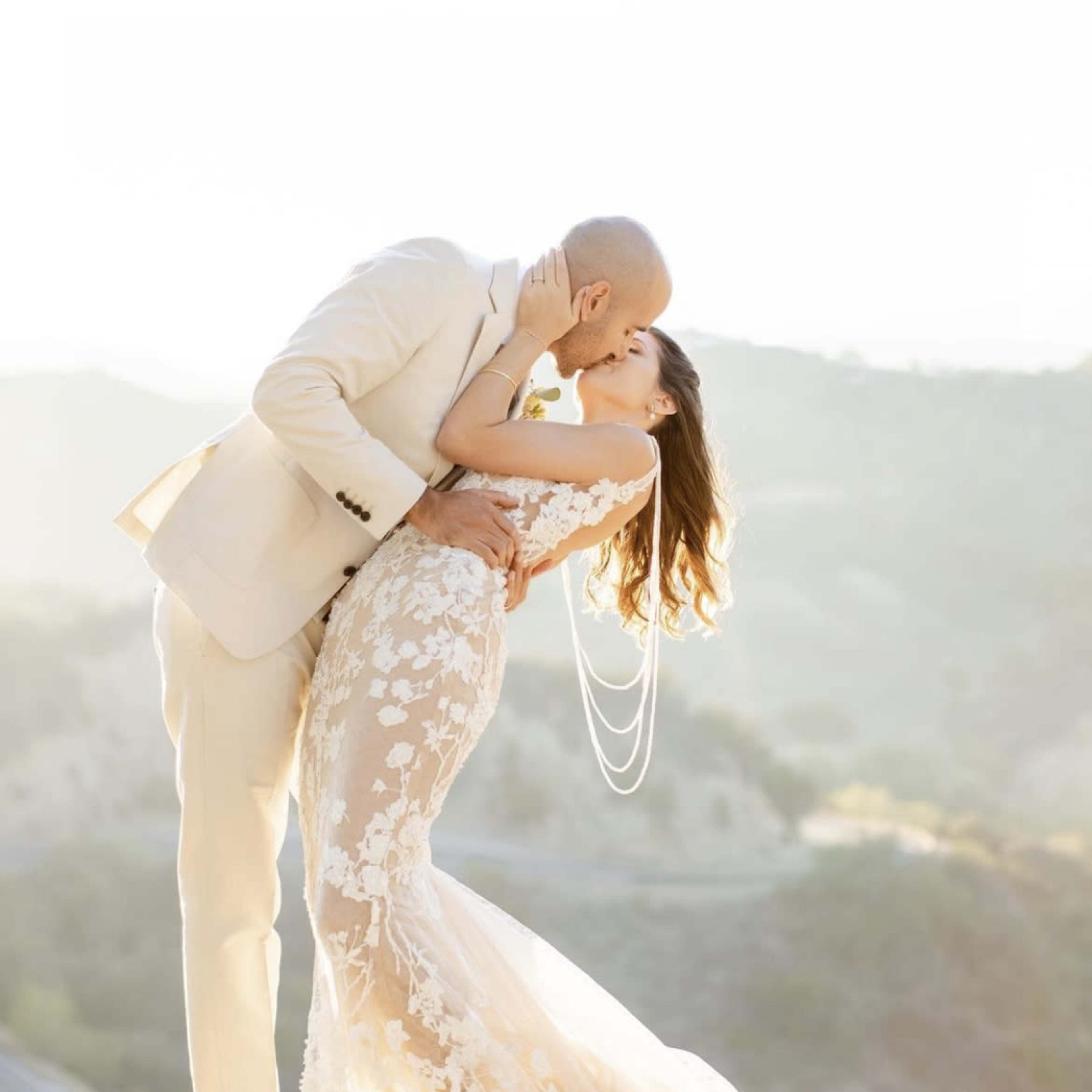 A couple kisses while standing on a hillside, dressed in wedding attire, with a scenic landscape in the background.