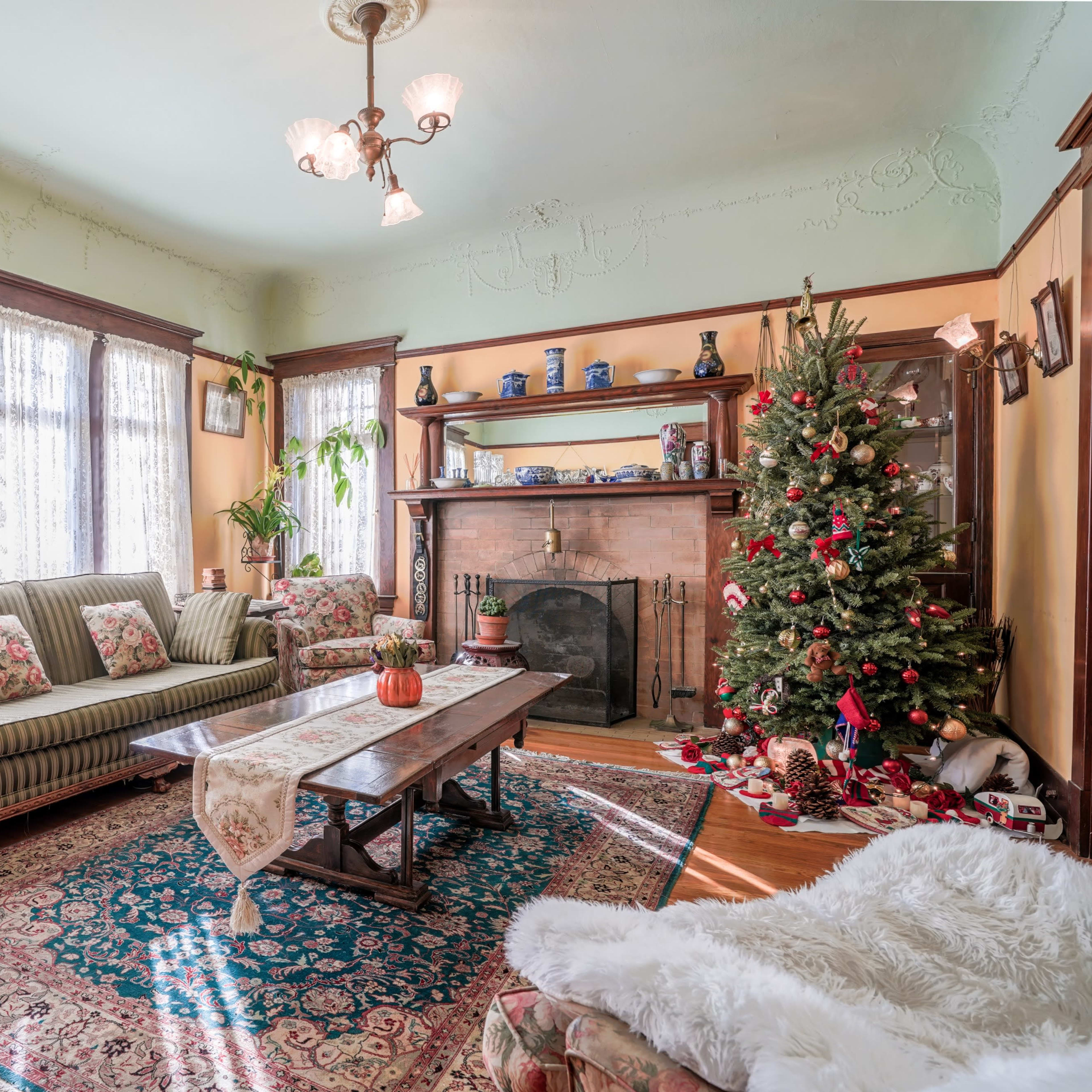A cozy living room features a patterned sofa, armchairs, a wooden coffee table, and a decorated Christmas tree beside a fireplace, with sunlight streaming through lace curtains.