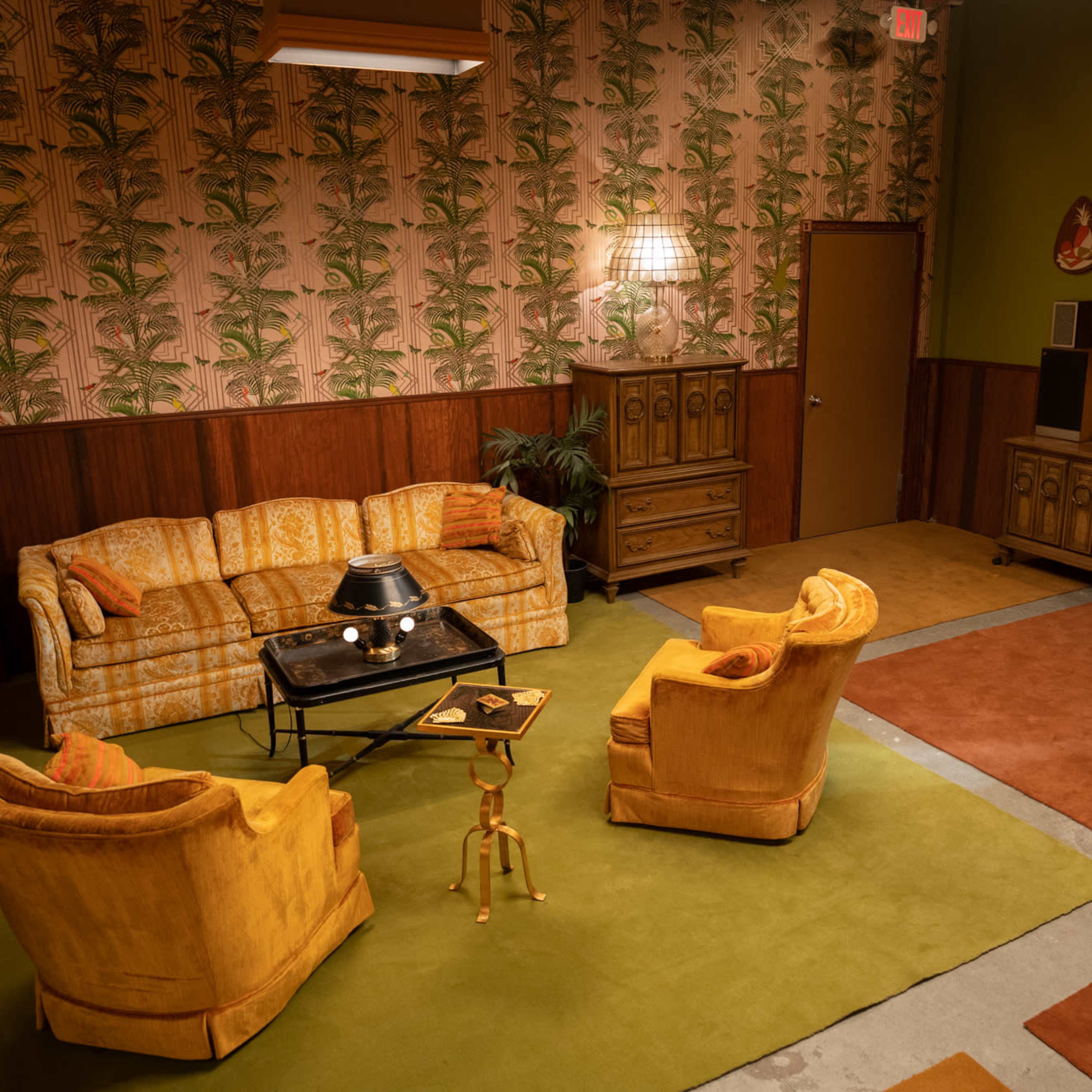 A vintage living room with a patterned wallpaper, yellow furniture, and a green carpet, featuring a coffee table and an entrance door.