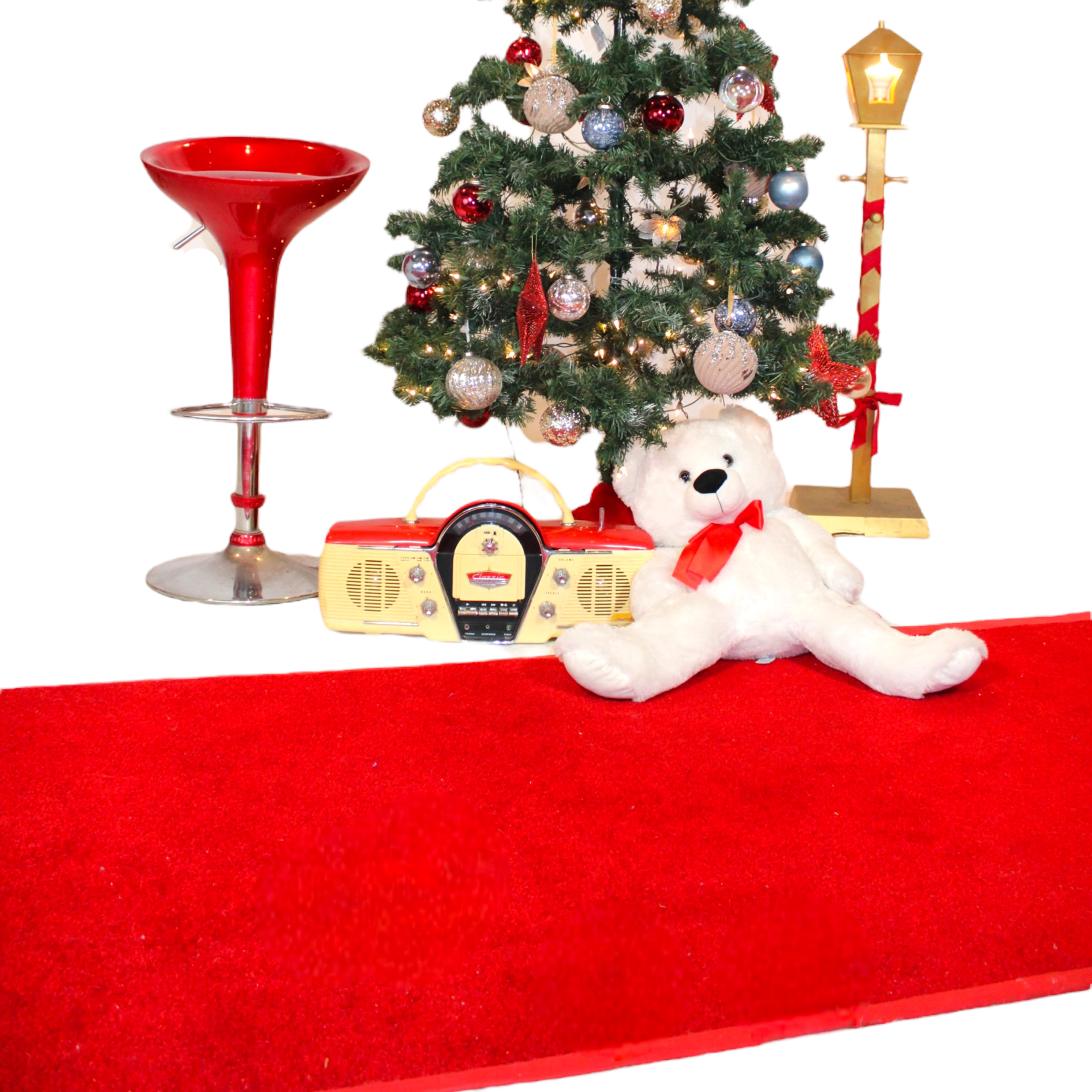 A Christmas tree adorned with ornaments stands beside a red carpet, a red bar stool, a lantern, and a plush teddy bear with a cassette player.