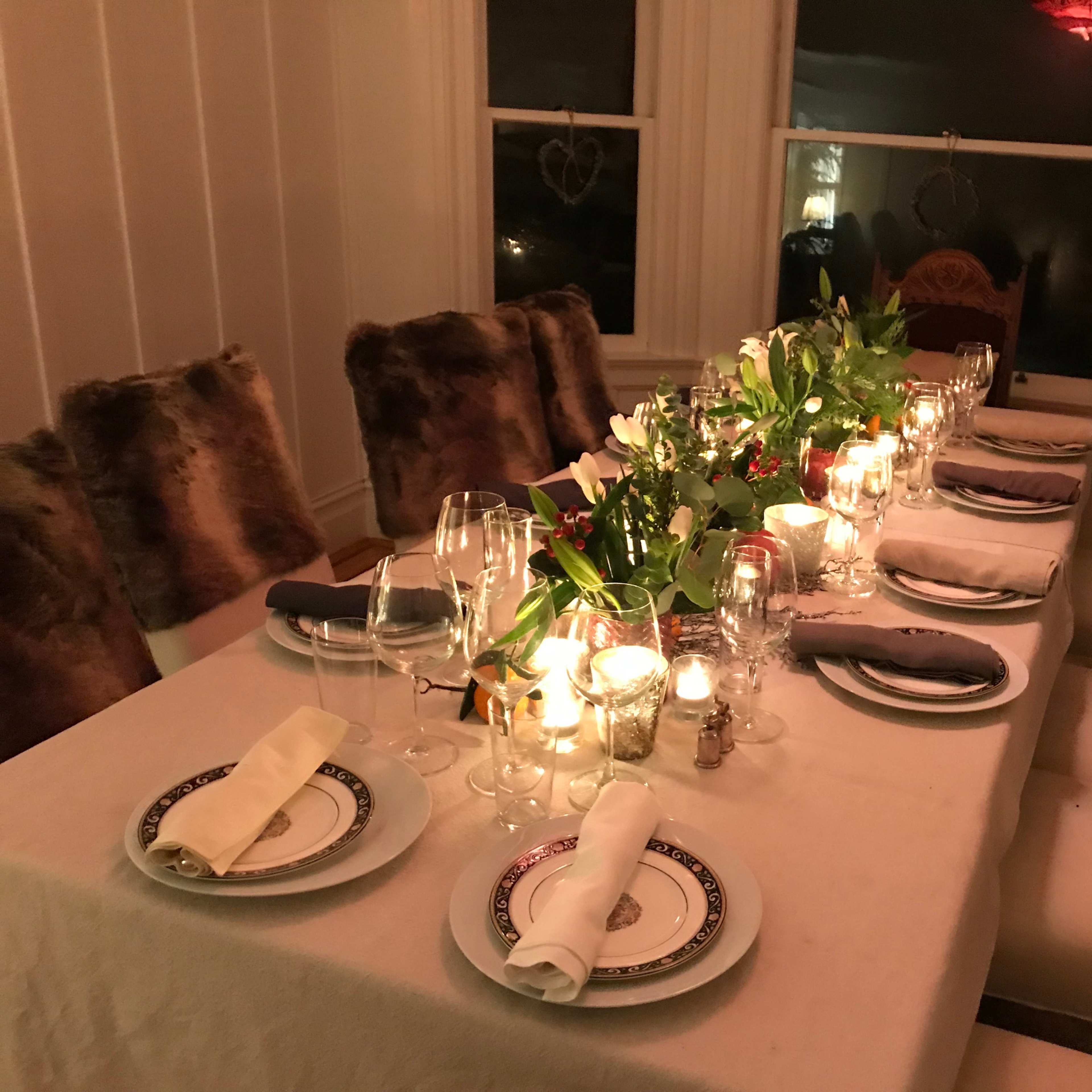 A long dining table is set with white tablecloths, plates, glassware, and candles, surrounded by chairs with fur covers.