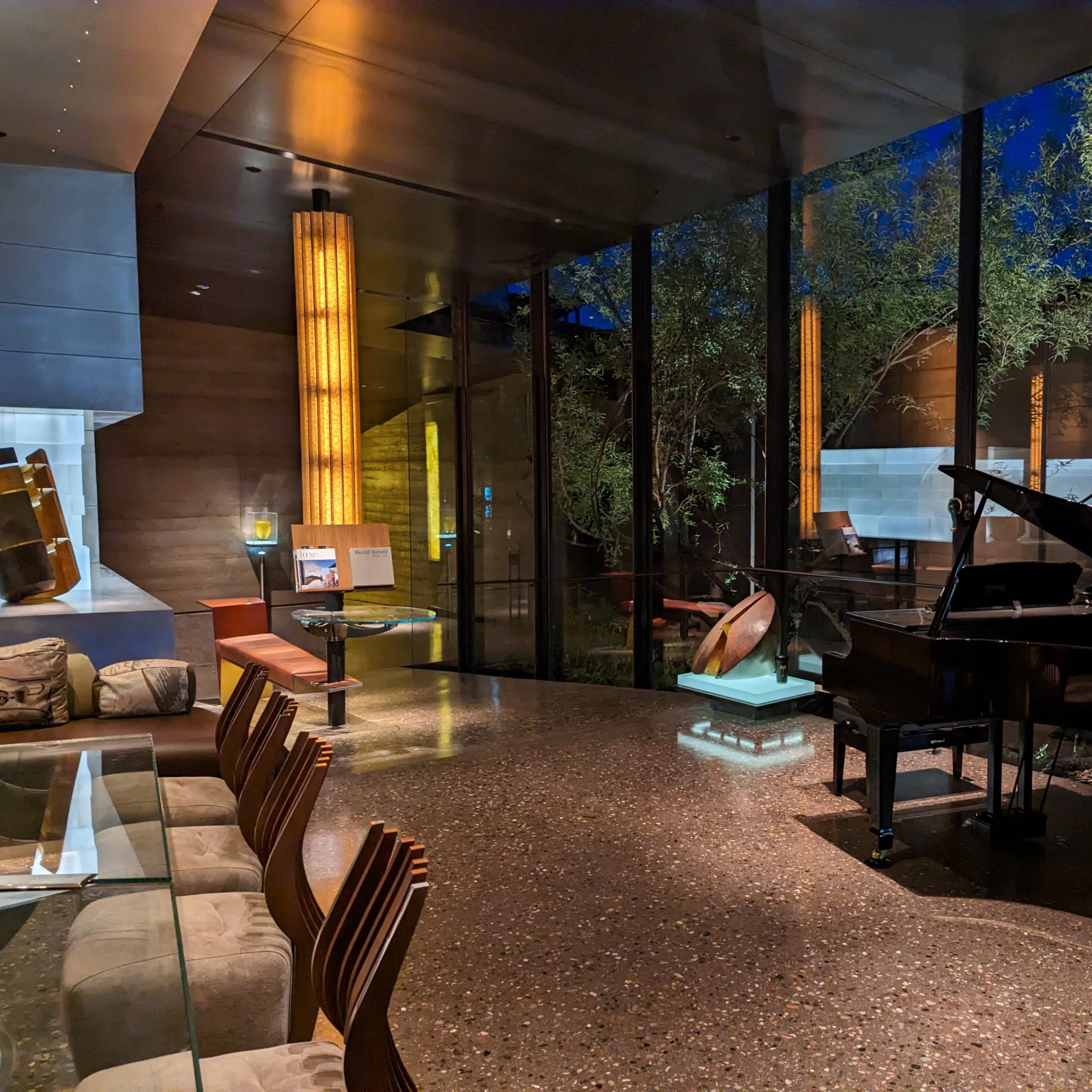 A grand piano sits in a dimly lit room with large glass windows overlooking a garden, accompanied by a modern dining area.