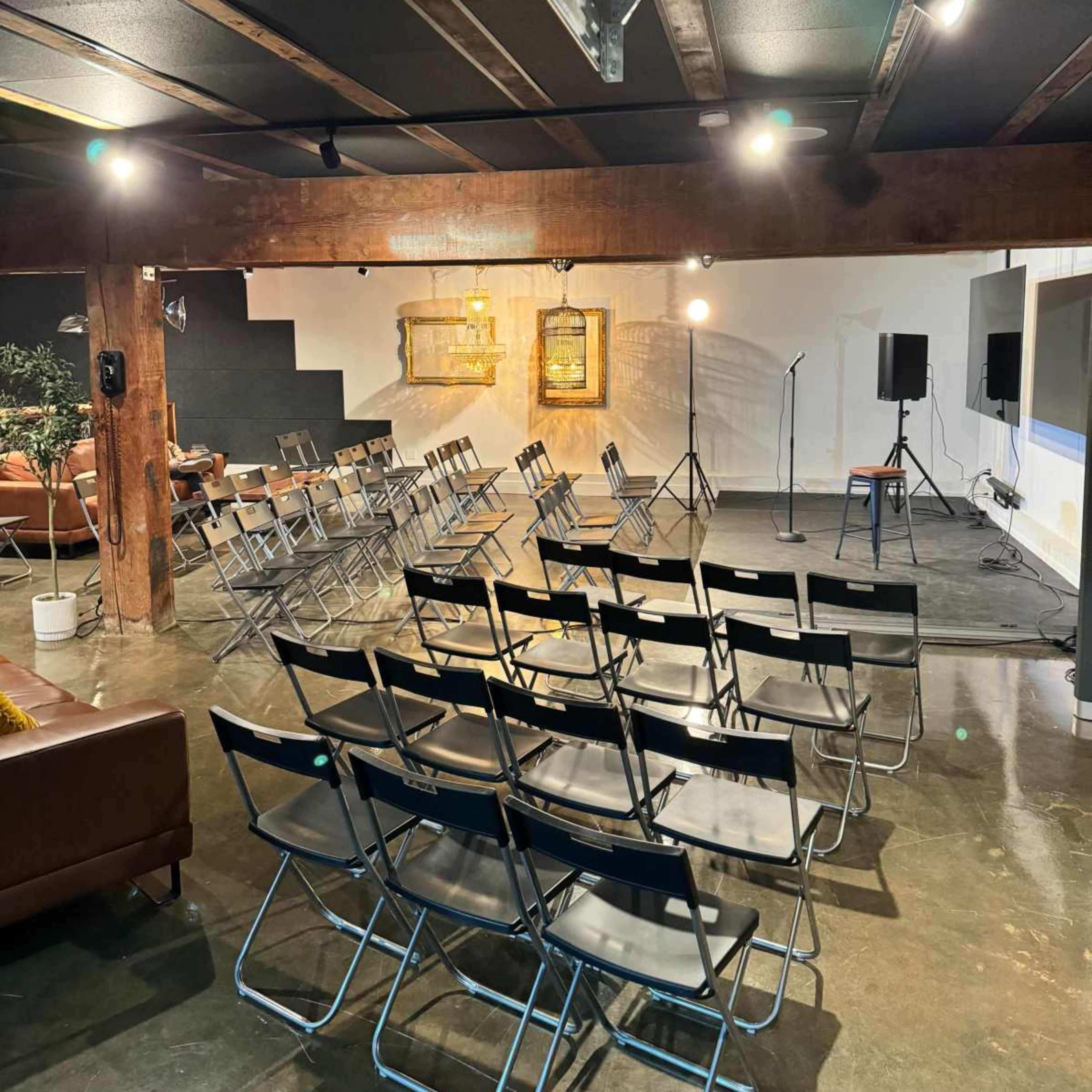 A spacious event room features rows of folding chairs facing a small stage with microphones and artwork on the walls.