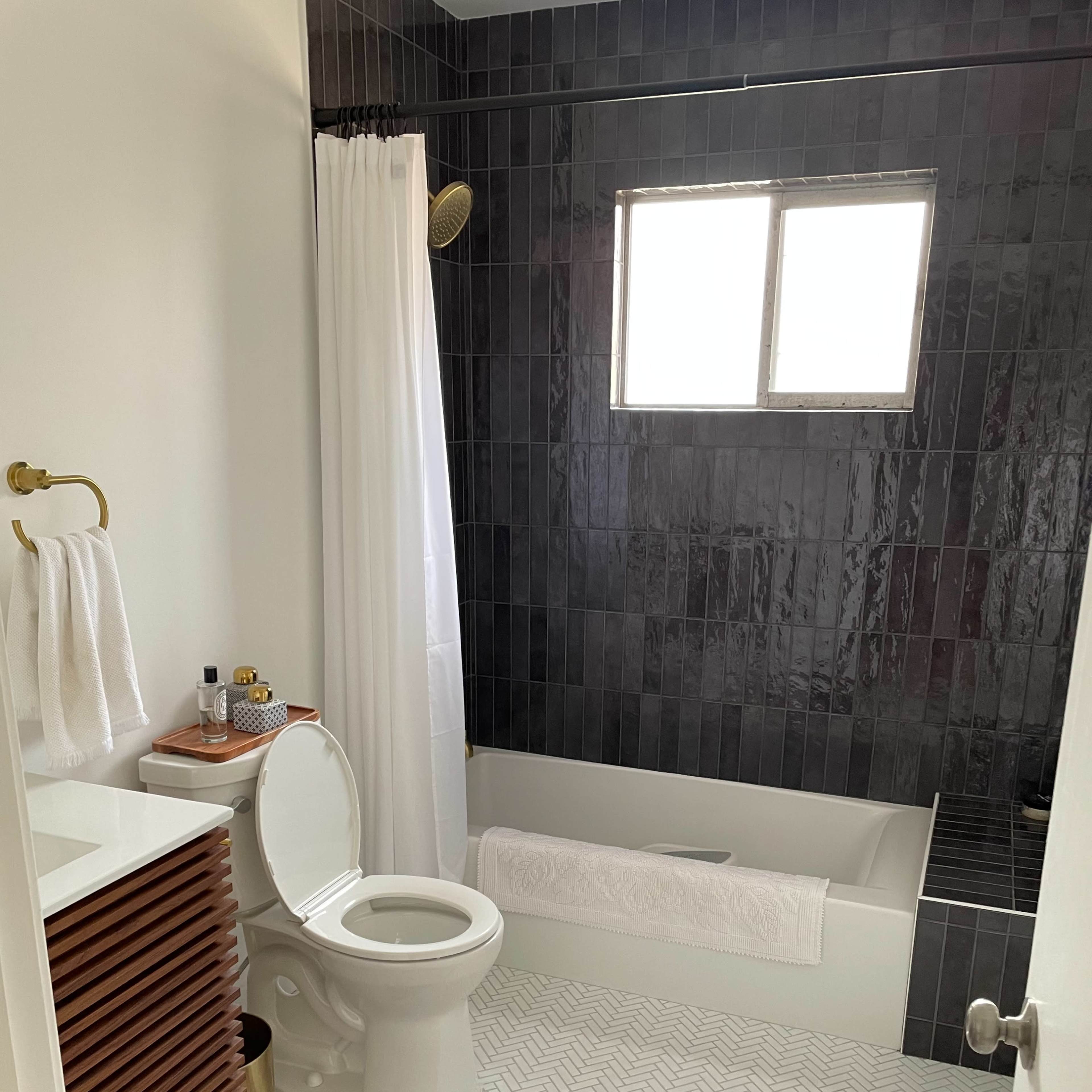A modern bathroom featuring a bathtub with a shower, a white toilet, a wooden vanity with a sink, and tiled black walls, along with a decorative rug on the floor.