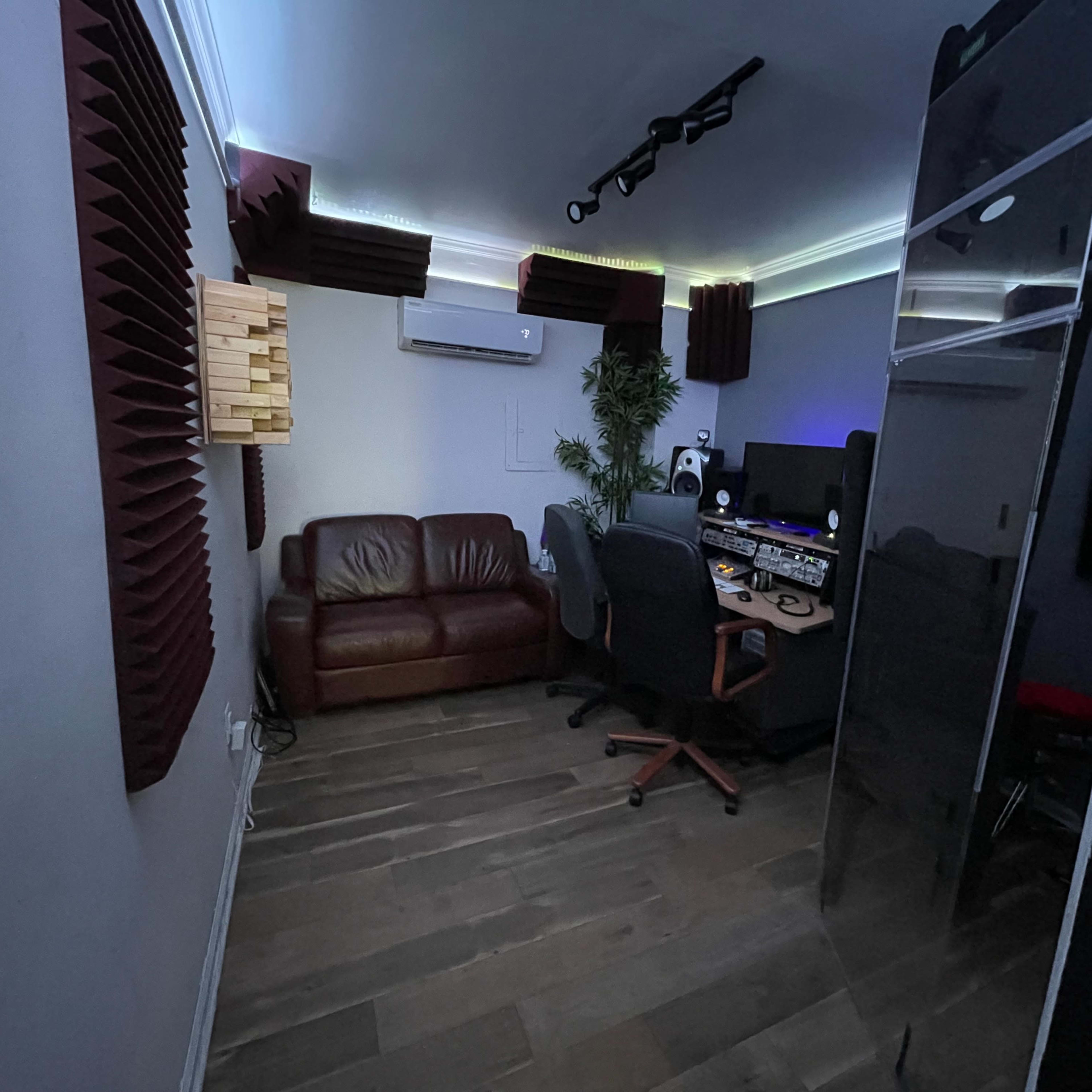 A small soundproof recording studio with a brown leather couch, a desk equipped with audio equipment, and acoustic panels on the walls.