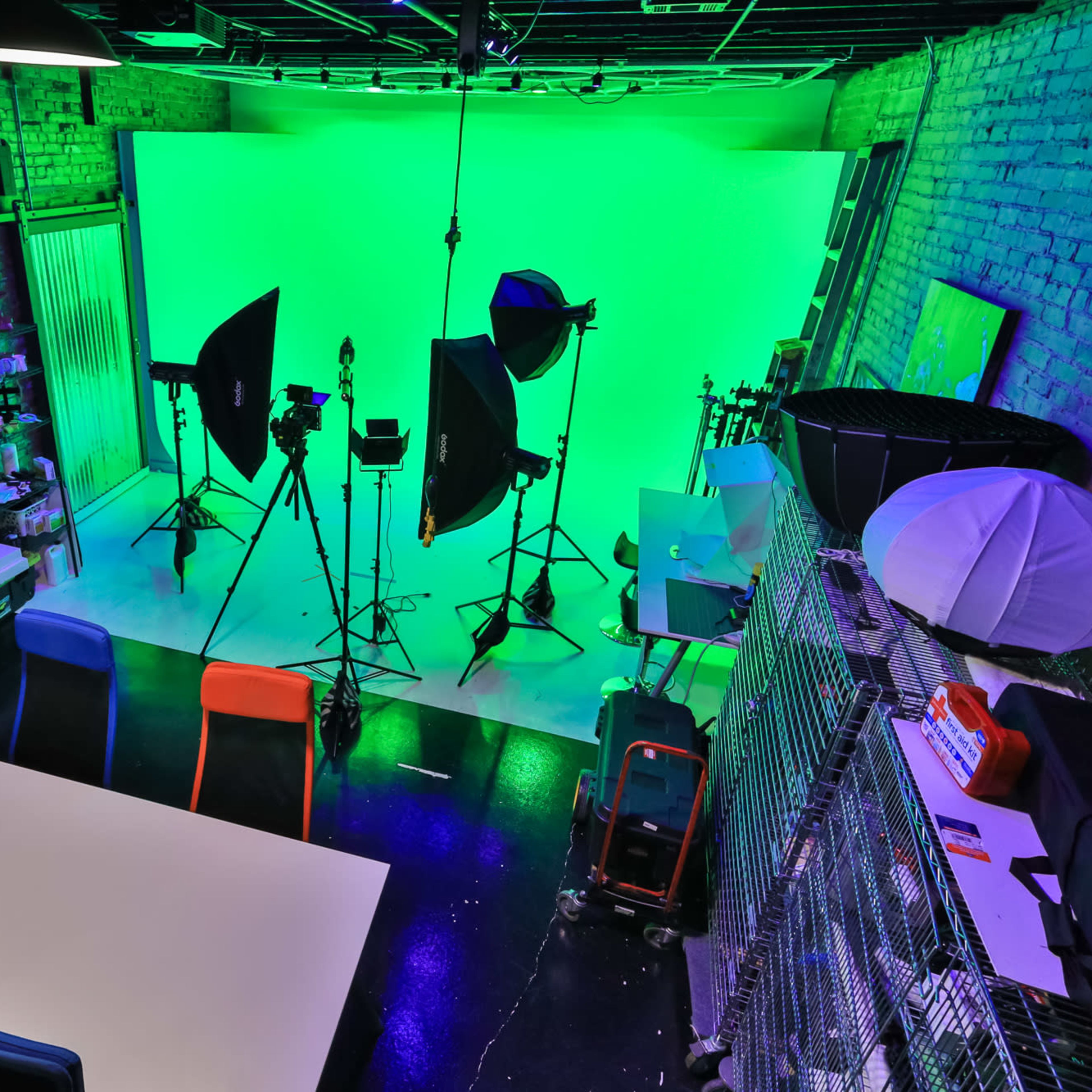 A brightly lit studio with a green screen backdrop, various lighting equipment, and a setup for video production, including a table and colorful chairs.
