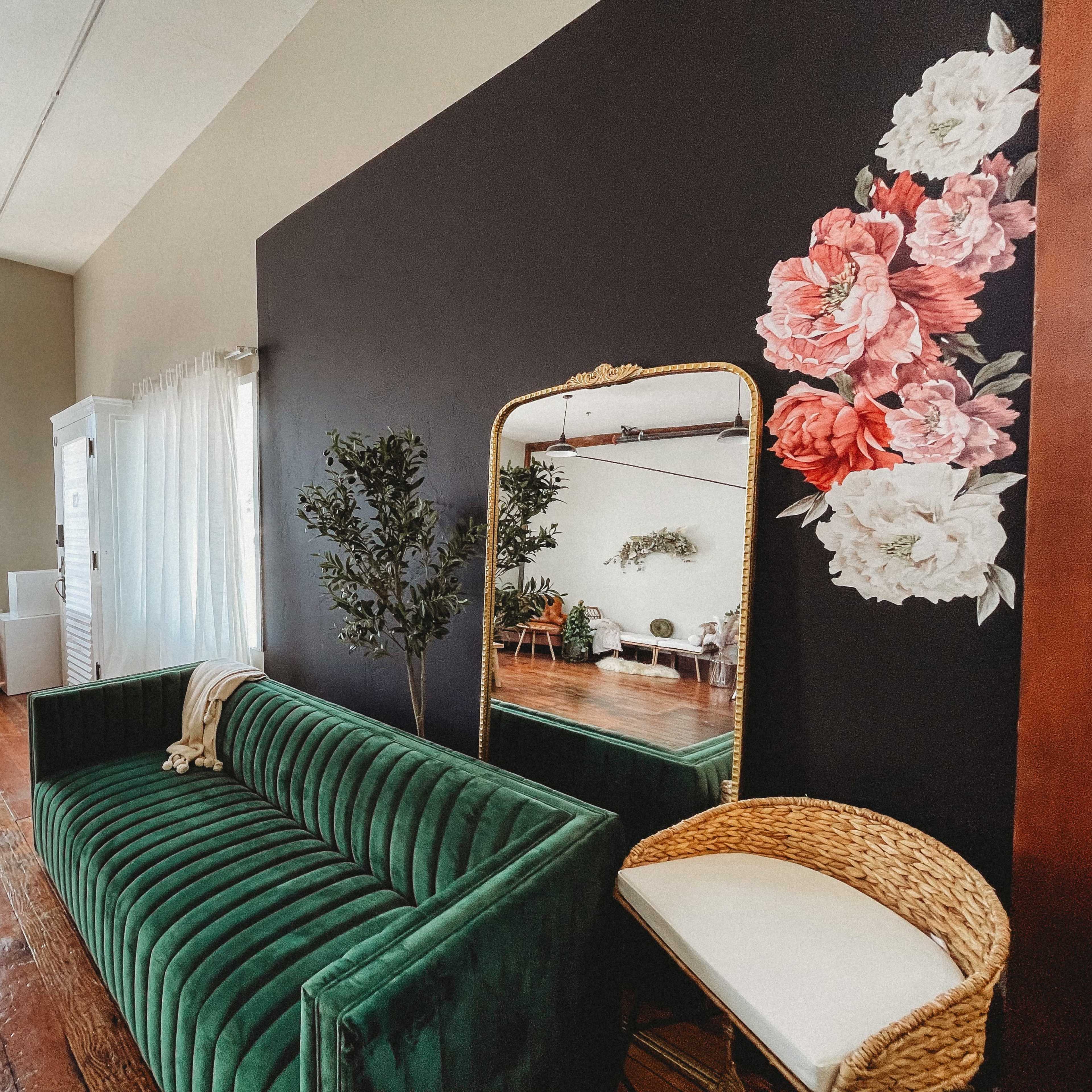 A stylish living room with a green velvet sofa, a large mirror, and floral wall art, complemented by plants and natural light.