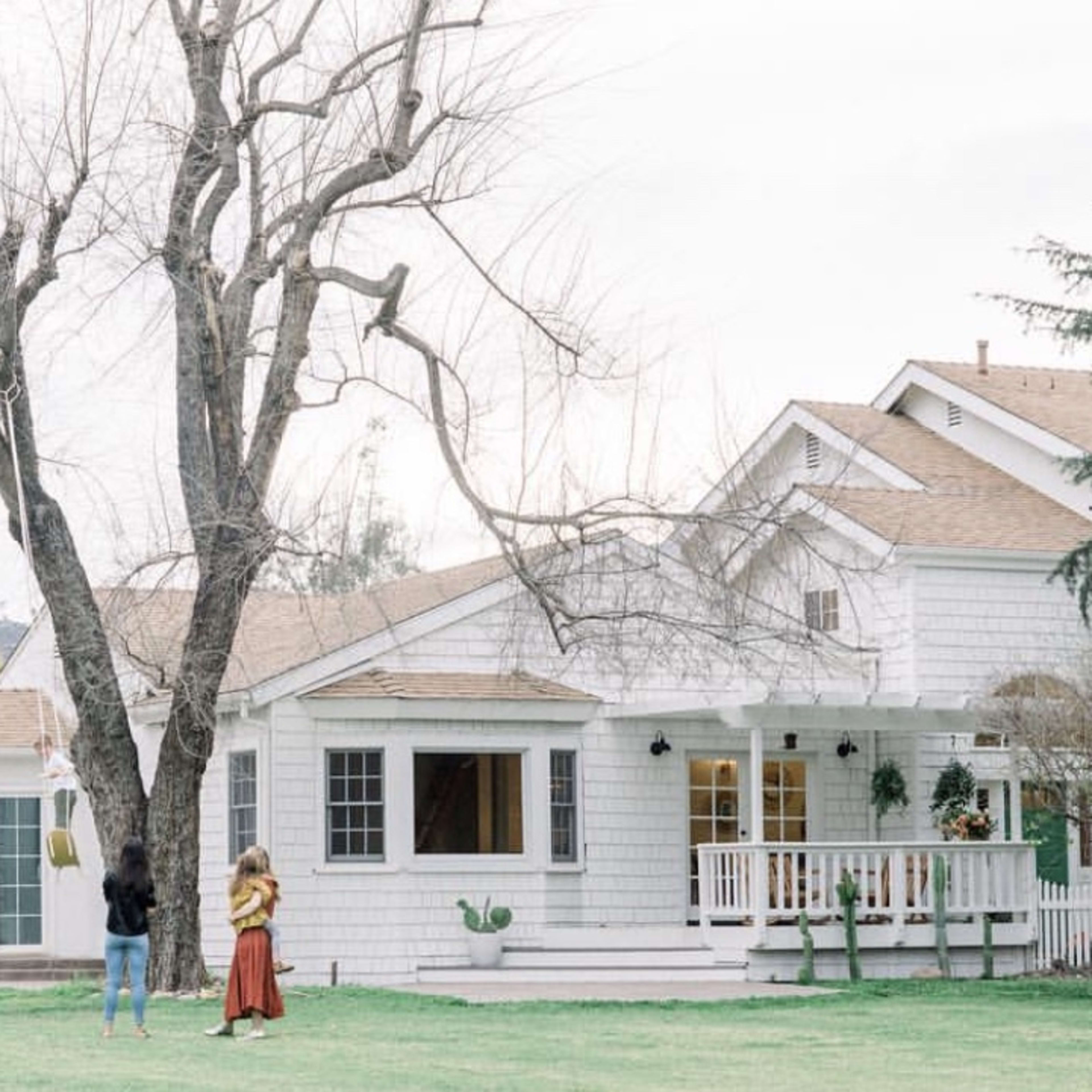 A white house with a porch and large windows sits in a yard where two people stand by a tree, and a dog is nearby.