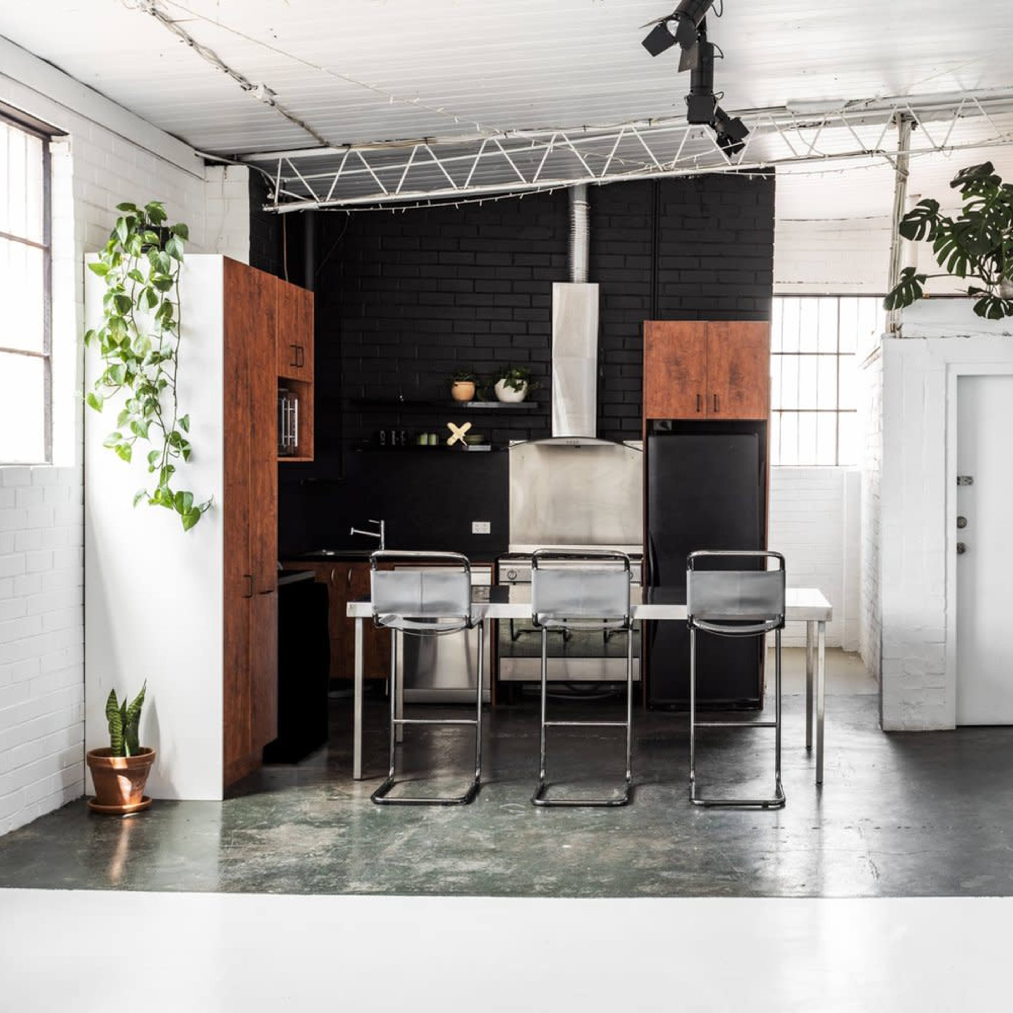A modern kitchen featuring a black and wooden cabinet setup, a stainless steel stove, and a dining table with bar stools, all surrounded by indoor plants.