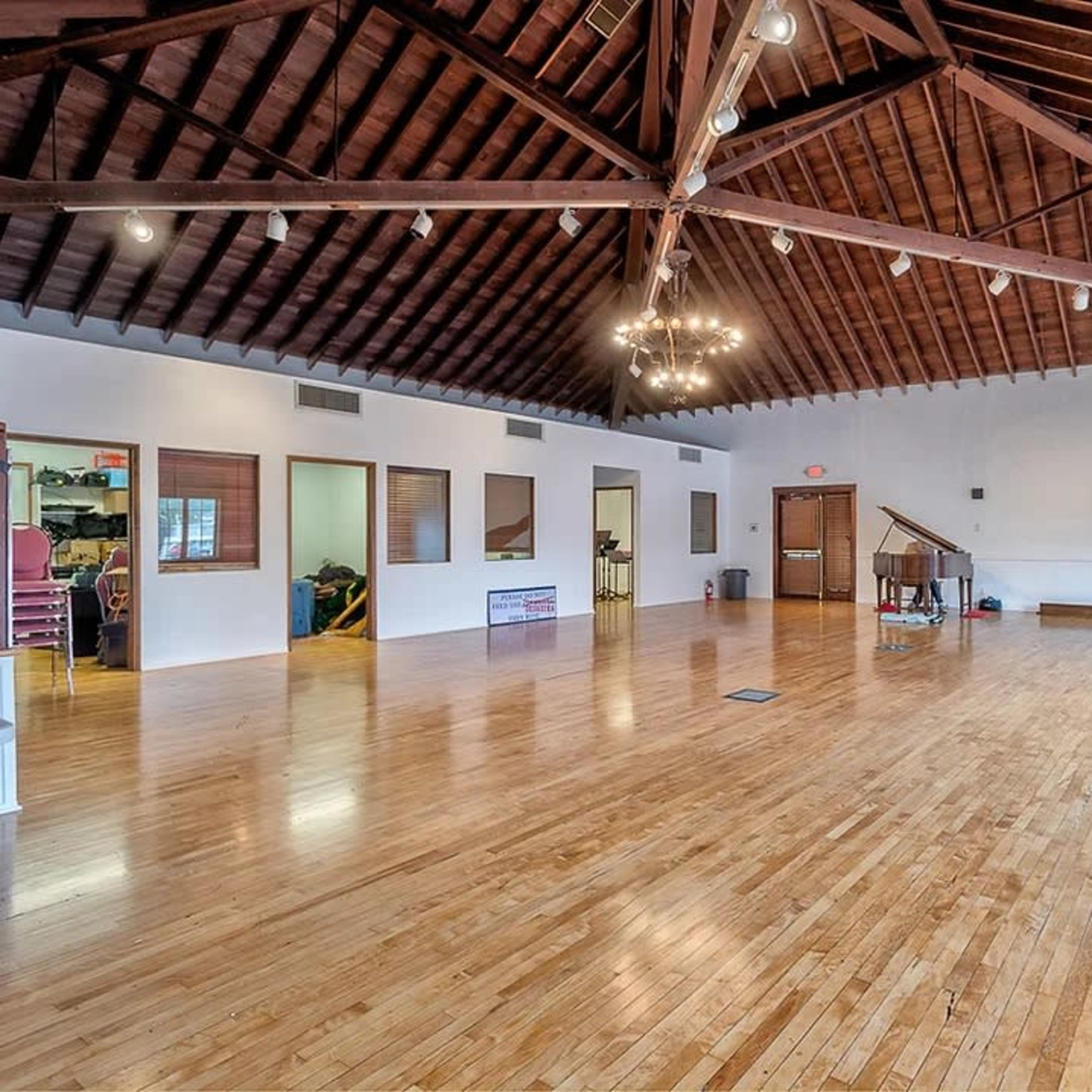 A spacious room with a wooden floor, high ceiling beams, and a piano against one wall.