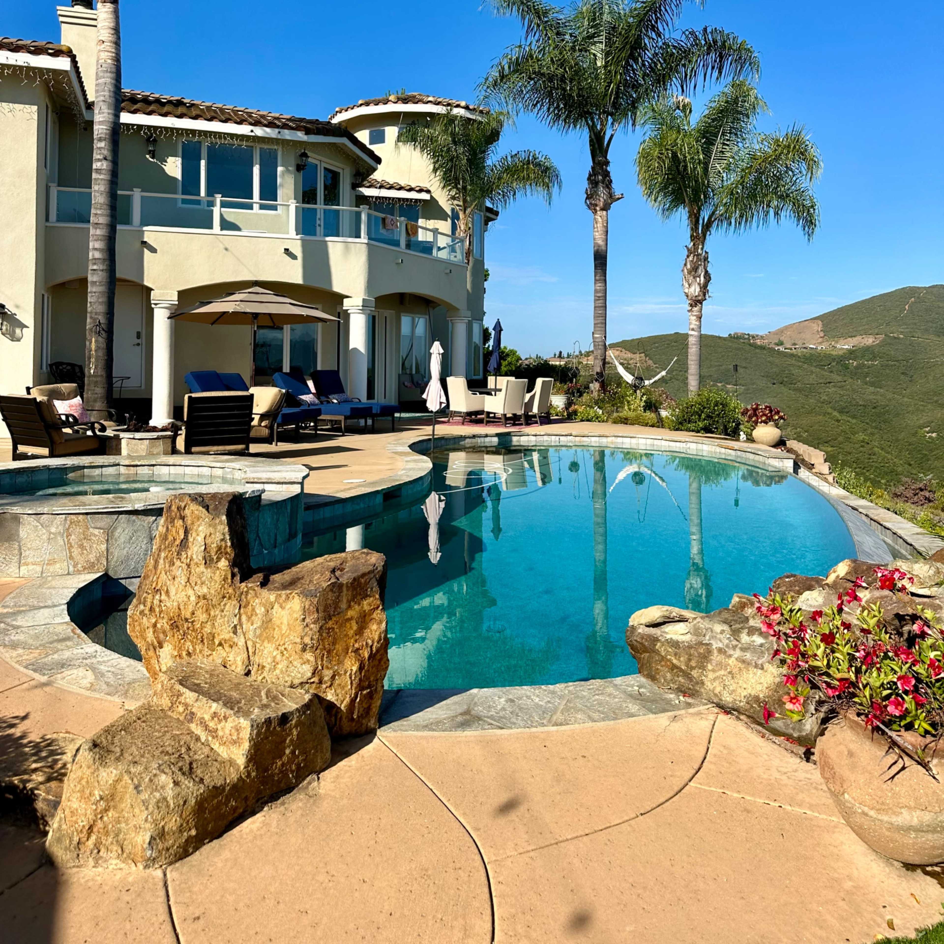 A luxurious home with a landscaped pool area is situated against a backdrop of rolling hills and clear blue skies.