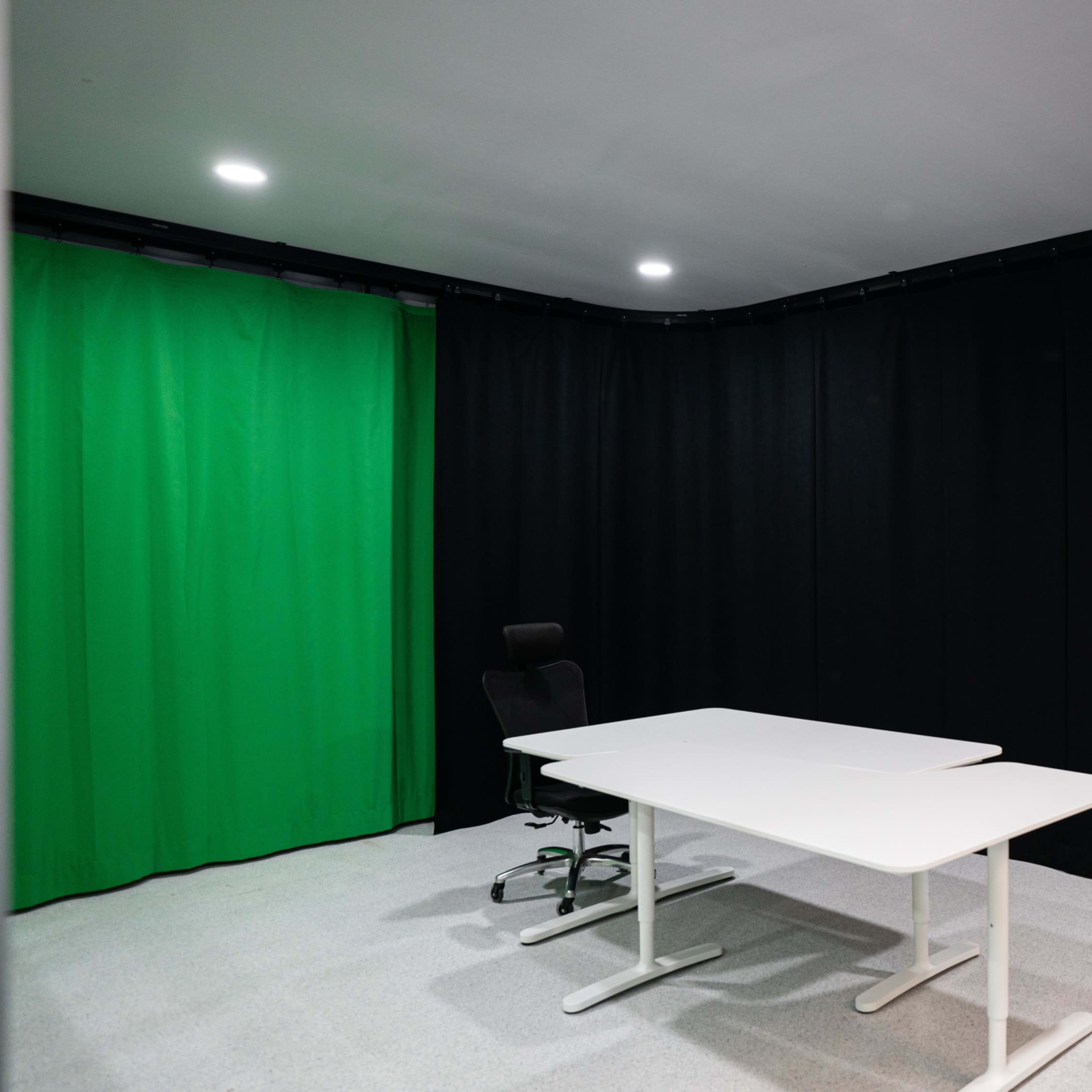 A small room features a green screen backdrop, a white table, a black chair, and a desk with various equipment.