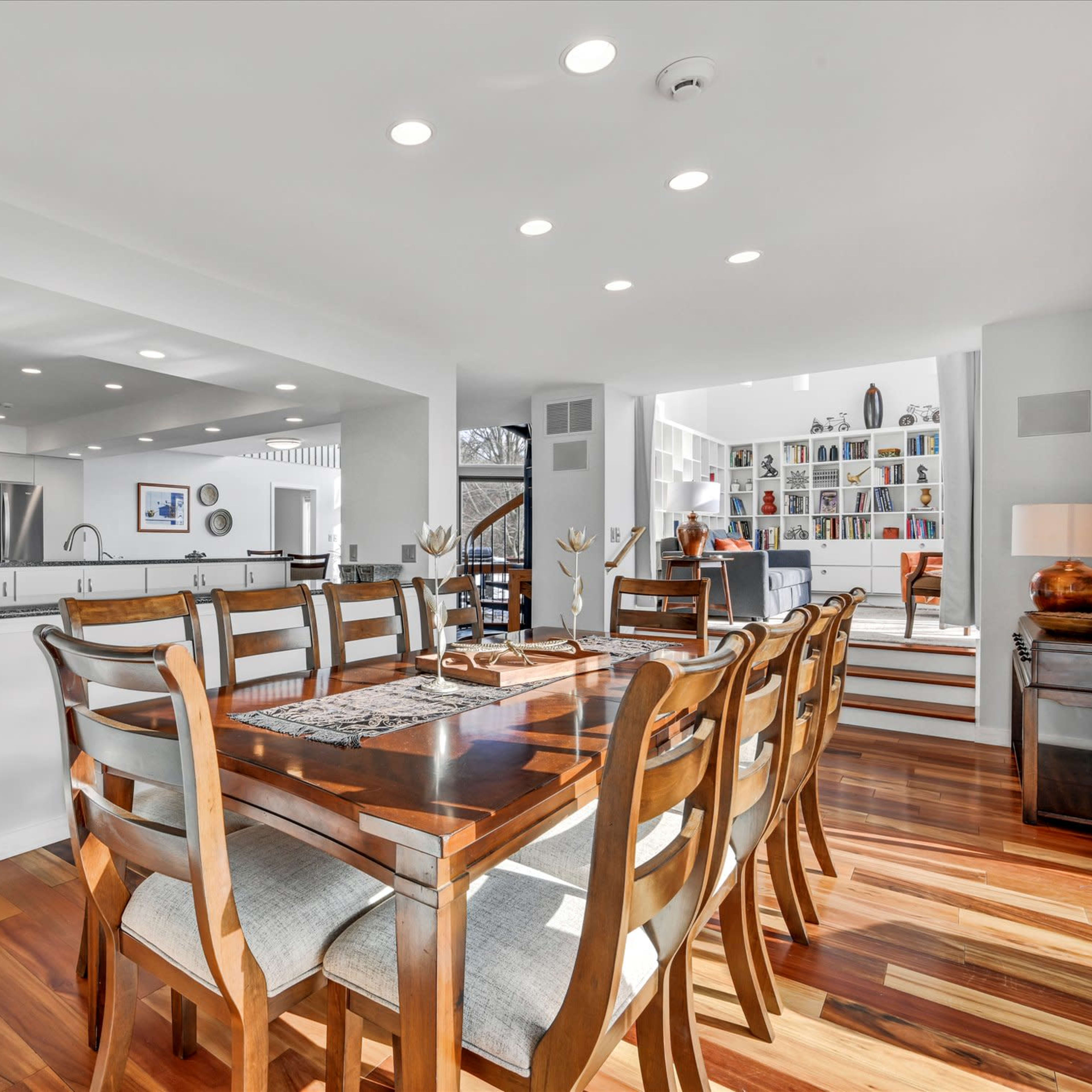 A modern dining area with a wooden table and chairs, leading to an open kitchen and living space filled with bookshelves.