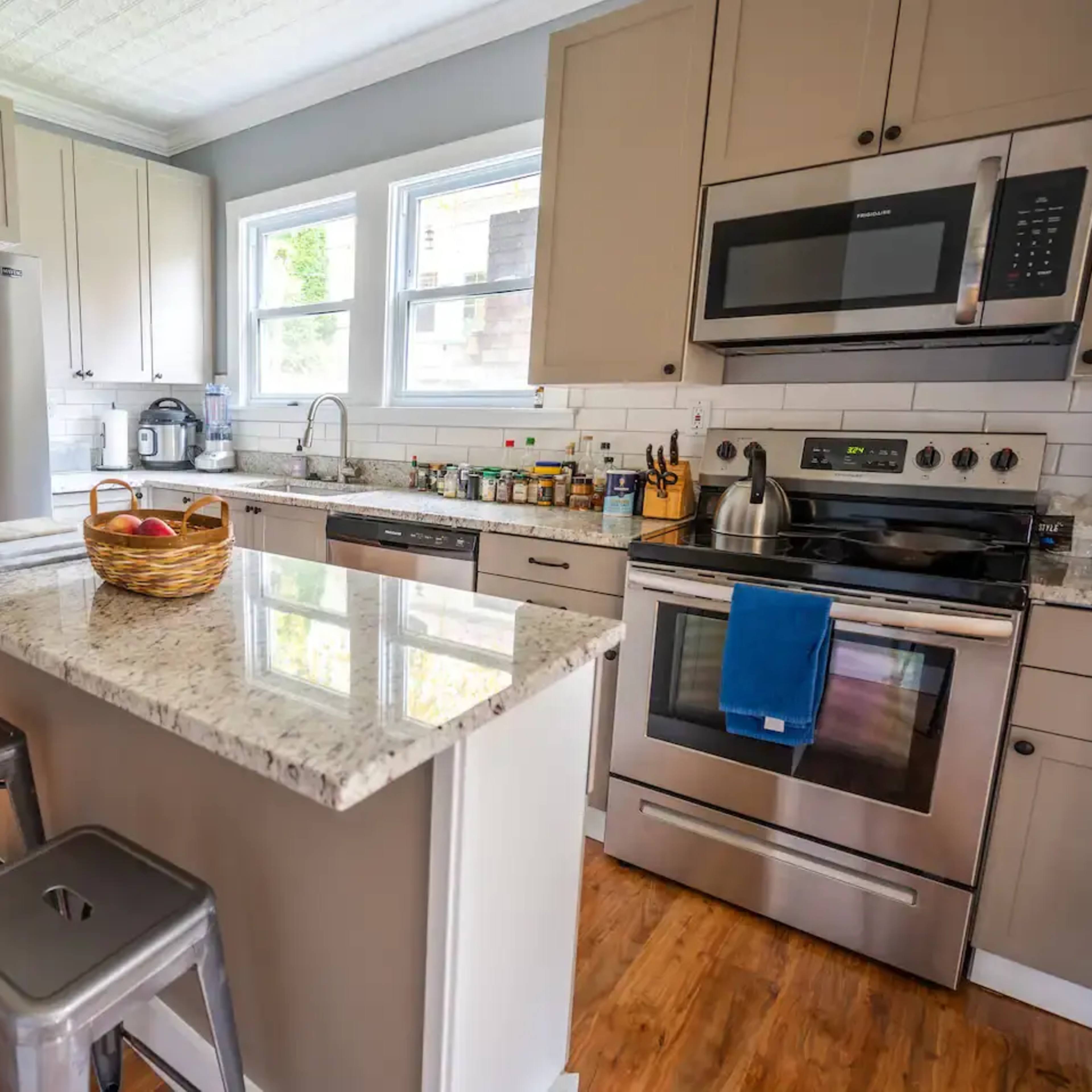 A modern kitchen features stainless steel appliances, a granite countertop island with a fruit bowl, and wooden flooring.