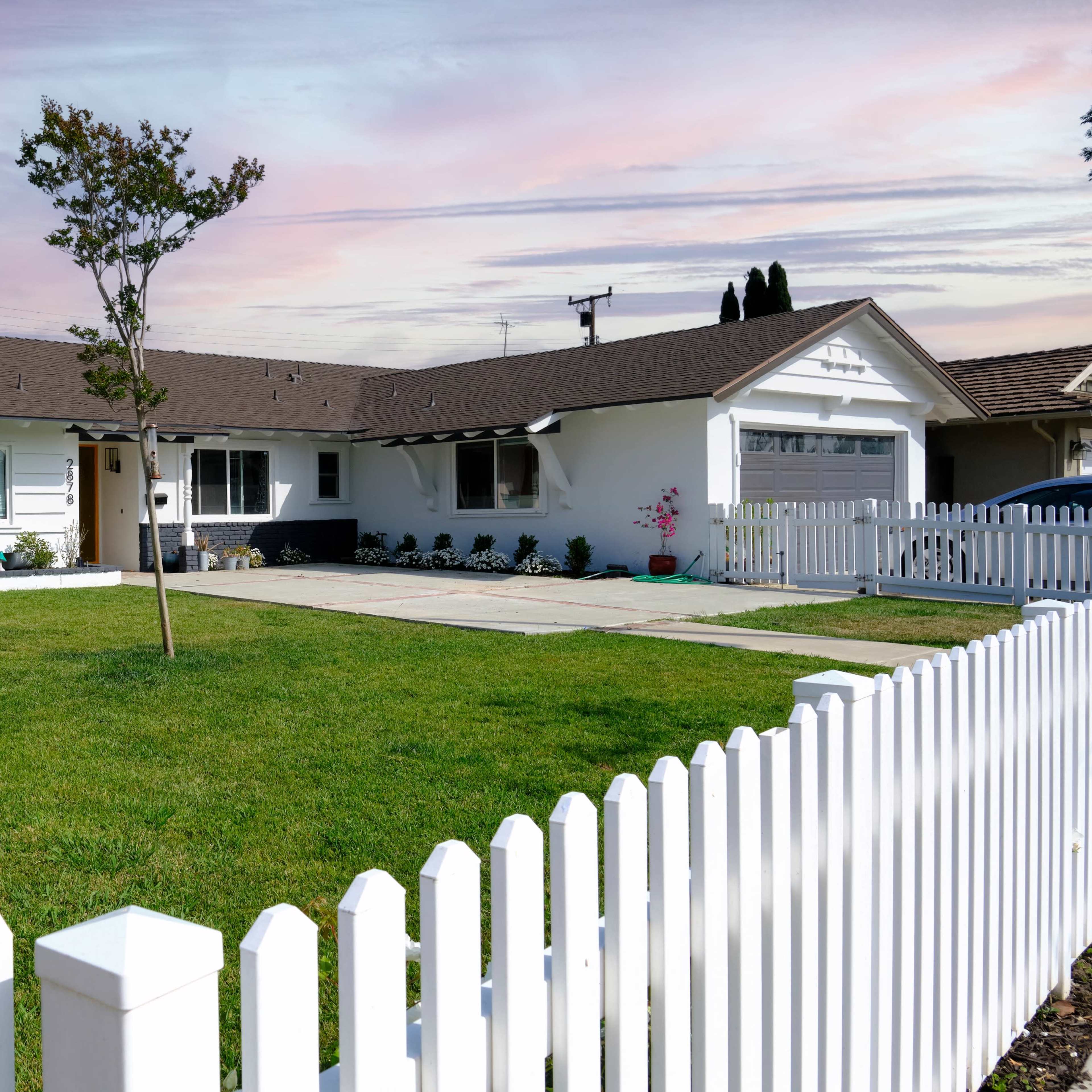 A single-story white house with a brown roof and a front yard surrounded by a white picket fence is shown.