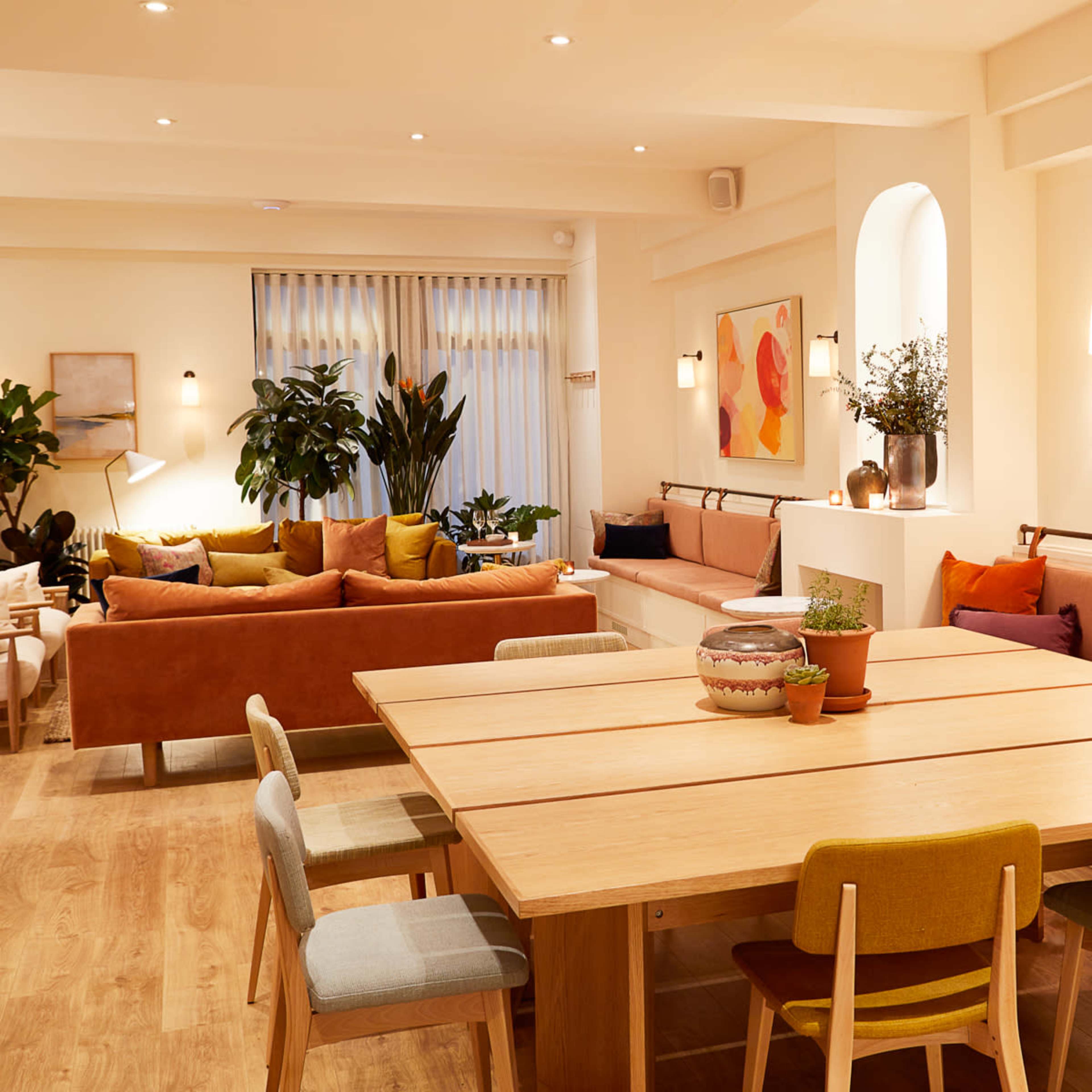 A modern interior space featuring a large wooden dining table surrounded by colorful chairs, with a lounge area equipped with sofas and plants.