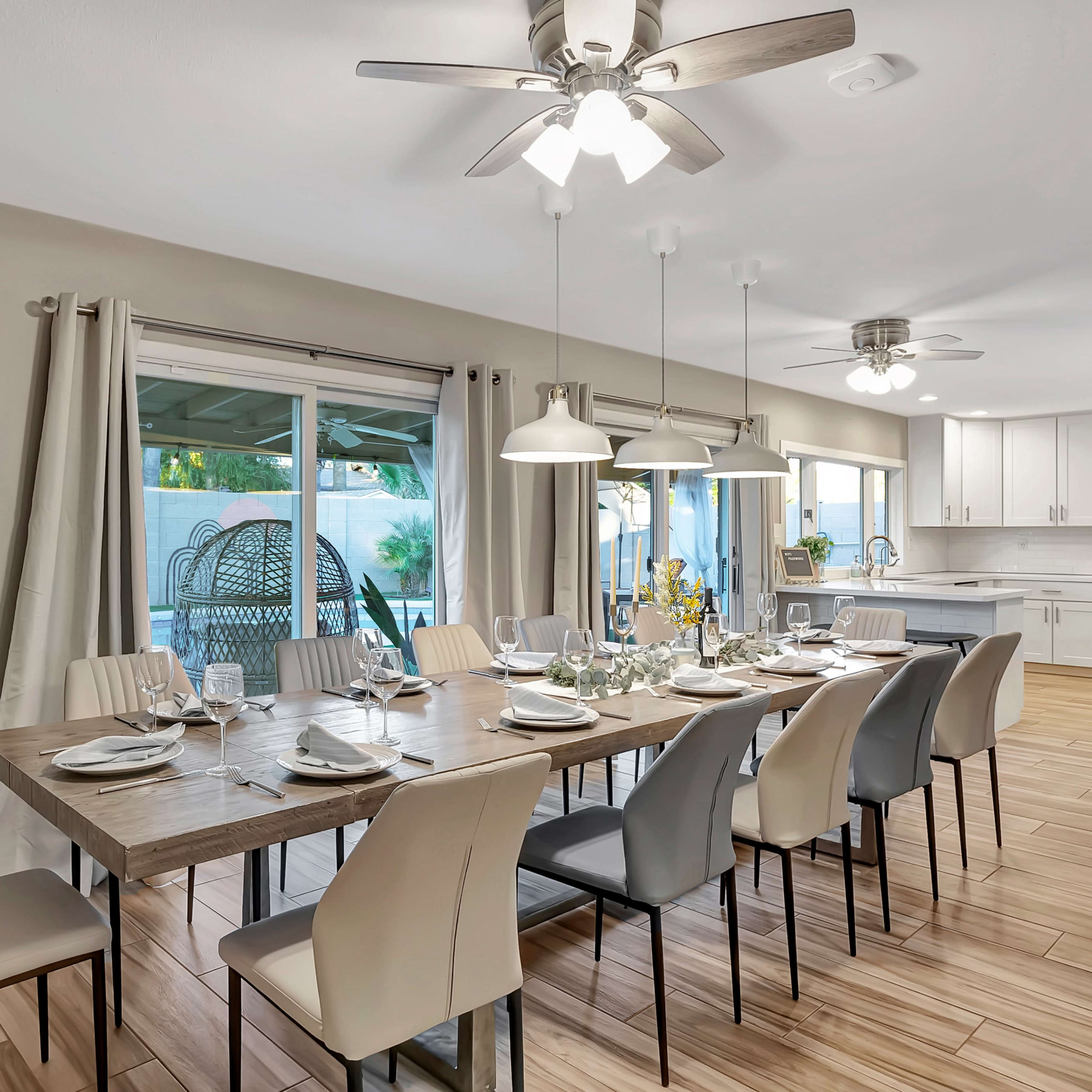 A modern dining room features a long wooden table set for dinner, surrounded by various chairs, with large windows overlooking a patio area.