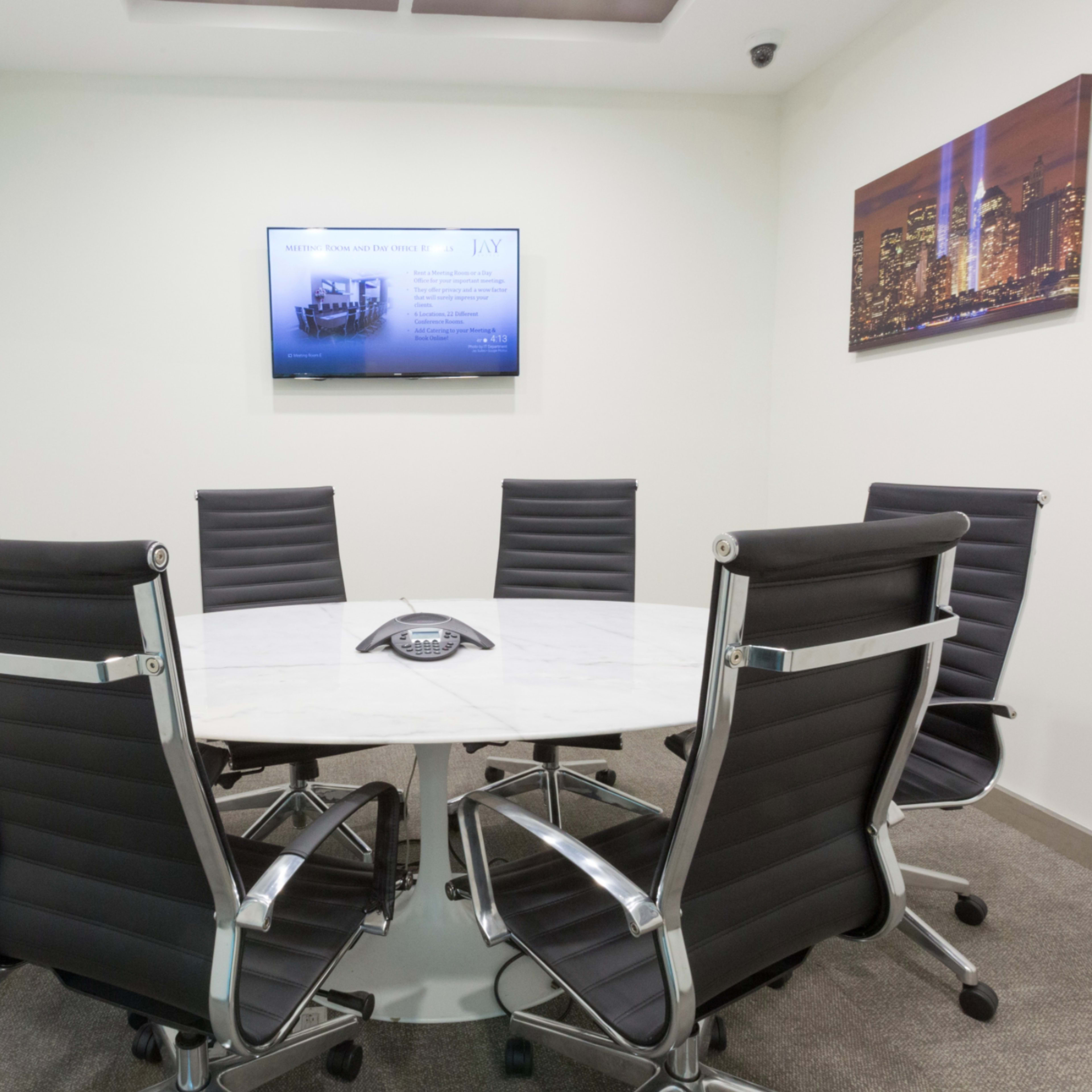 A modern conference room features a round marble table surrounded by six black chairs, with a wall-mounted screen displaying a presentation.