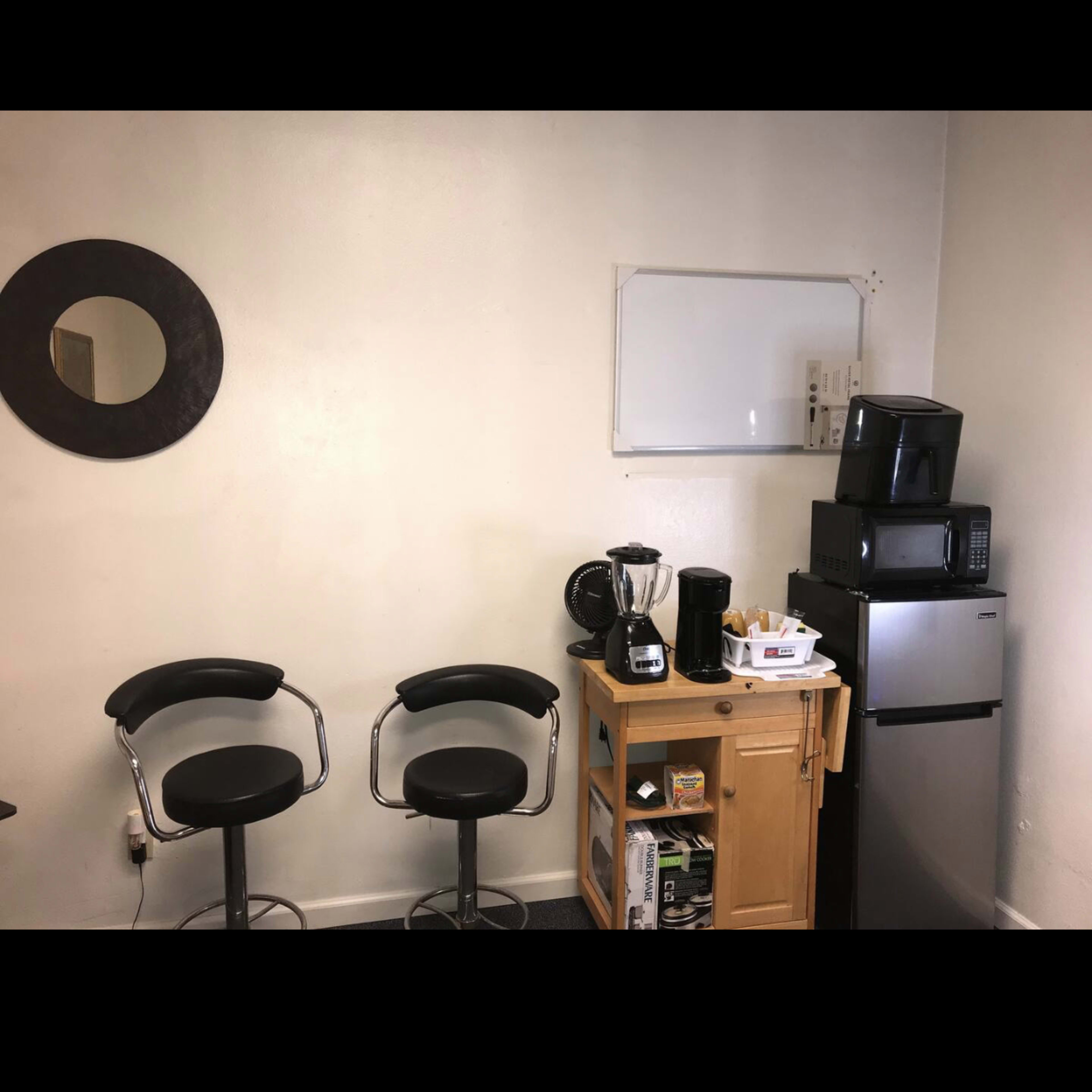A small kitchenette with two black bar stools, a wooden cart with kitchen appliances, and a refrigerator against a light-colored wall.