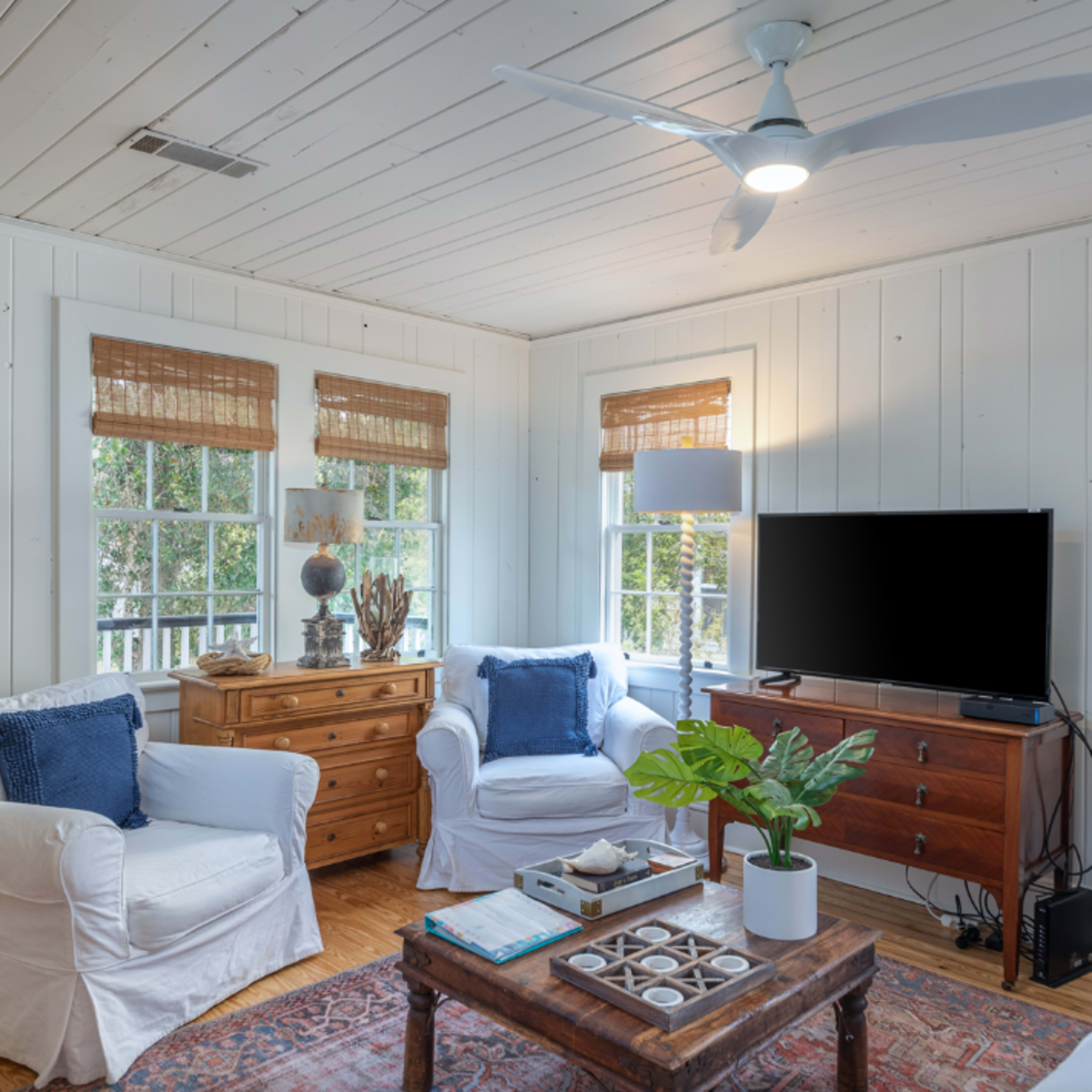 A cozy living room with white walls, a wooden floor, two windows with blinds, and a television on a wooden media console.