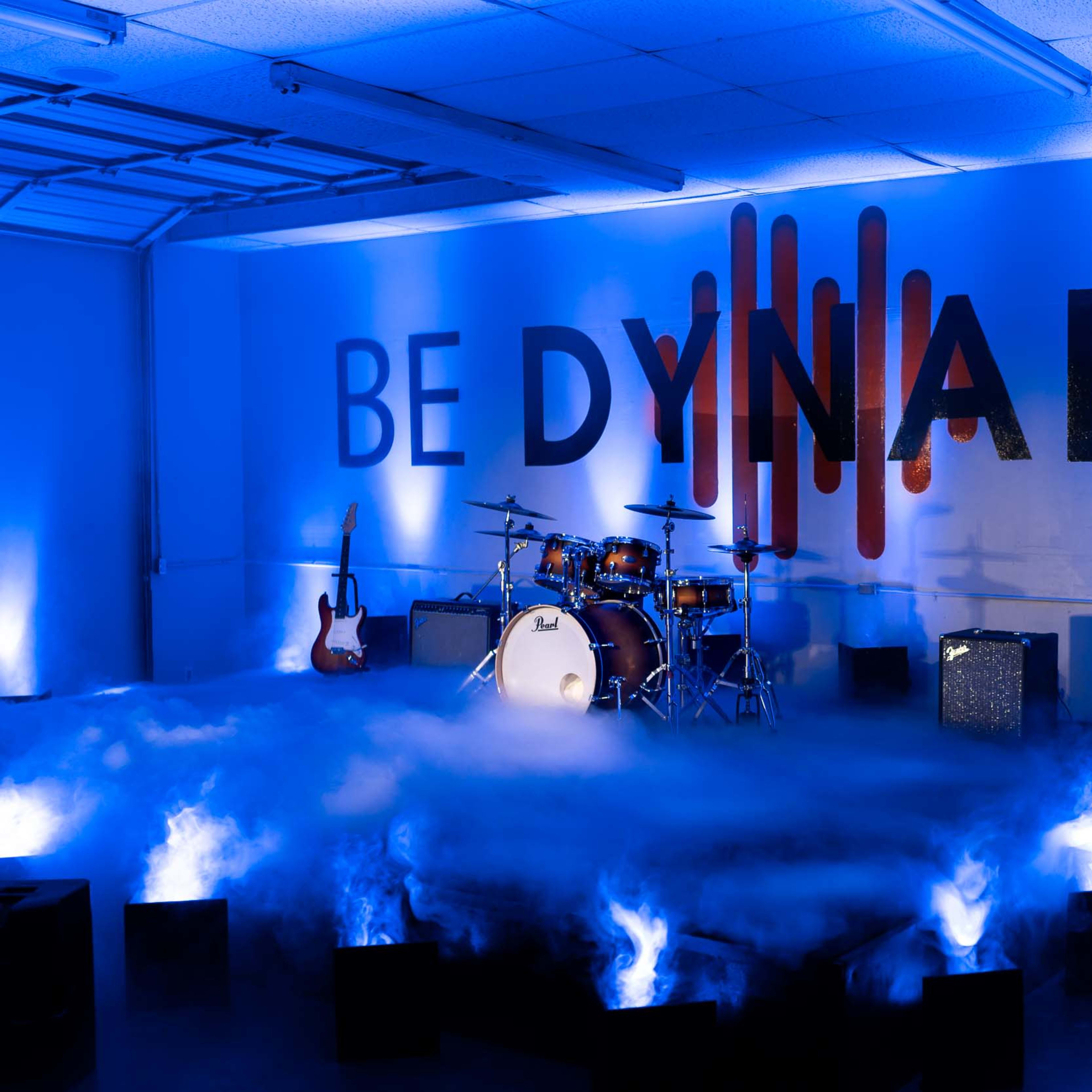 A music setup featuring a drum kit and guitar against a wall with the phrase "BE DYNAMIC," surrounded by smoke and illuminated by blue lights.