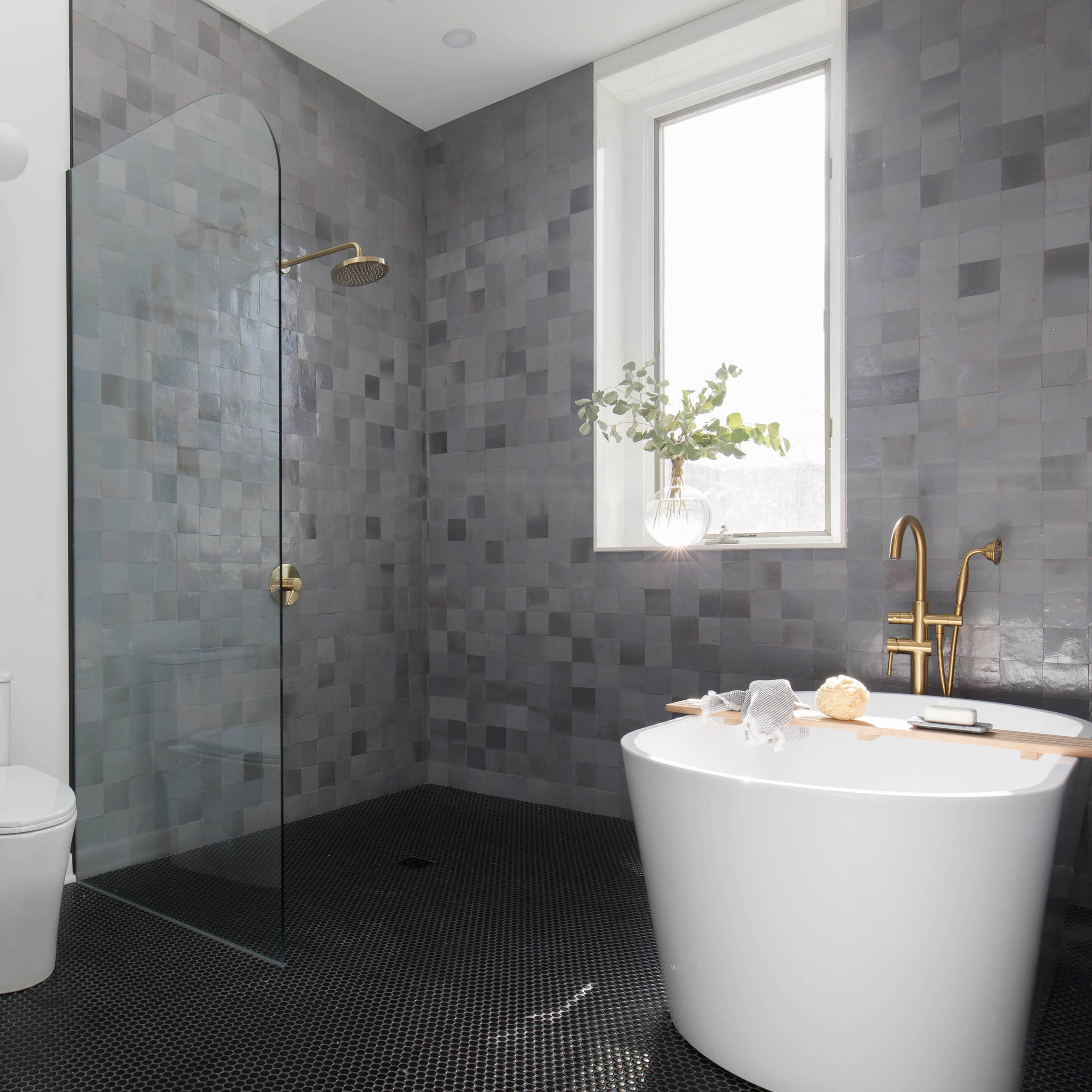 A modern bathroom featuring a glass shower, a freestanding white bathtub, a toilet, and a gray tiled wall with a small window.