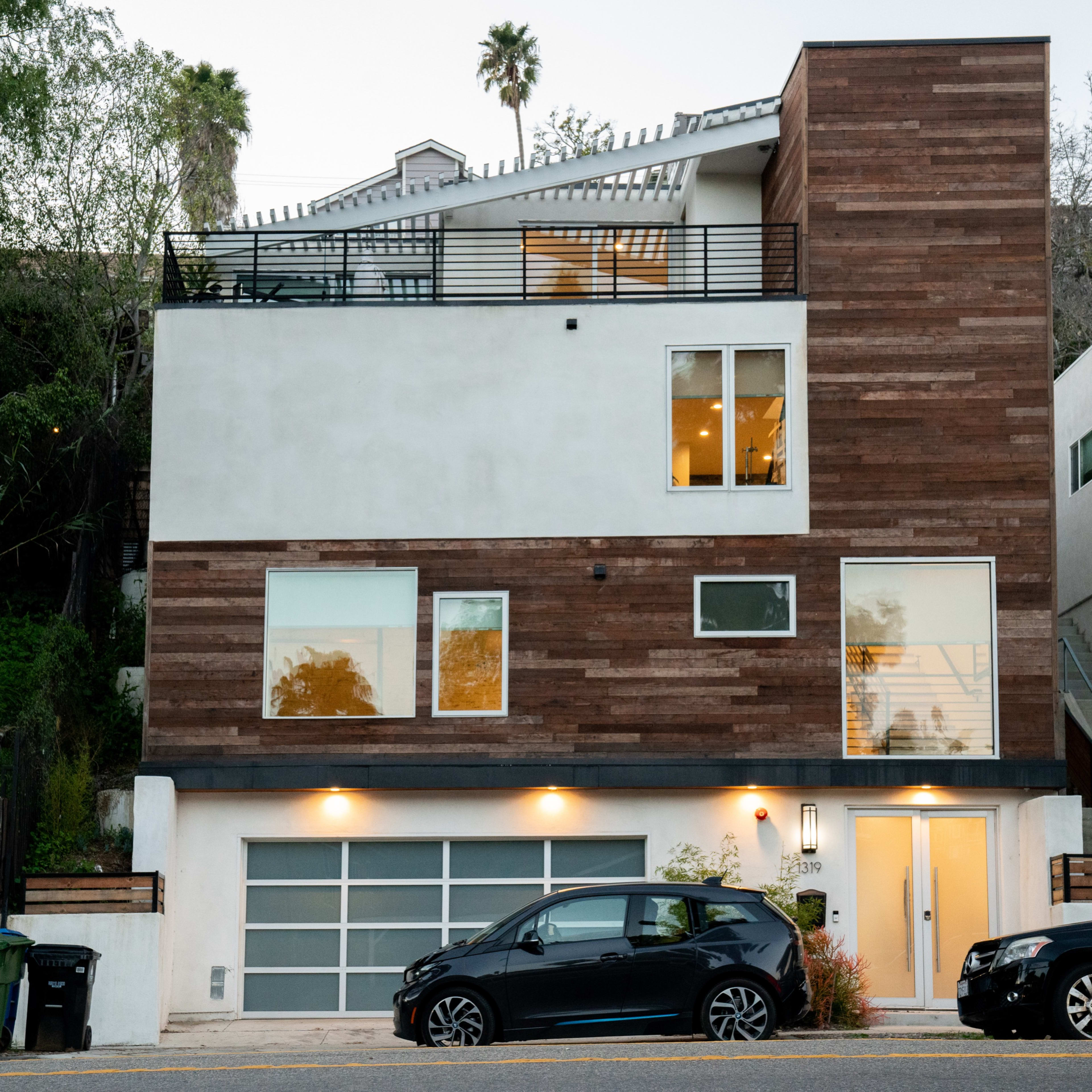 A modern two-story house with a wooden facade and large windows, located on a hillside street with parked cars in front.