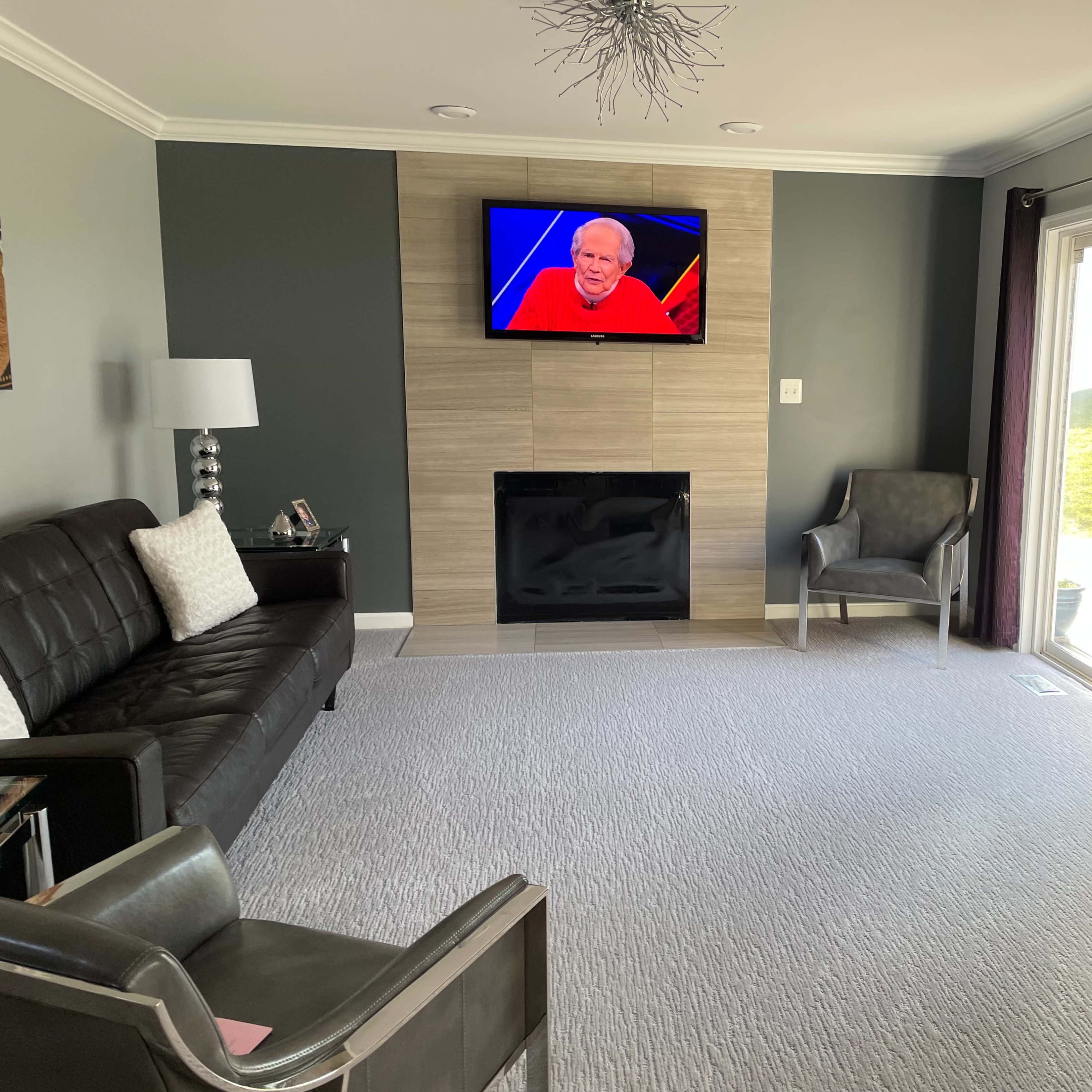 A living room with a dark couch, two chairs, a television mounted on the wall, and a large window leading to an outside view.