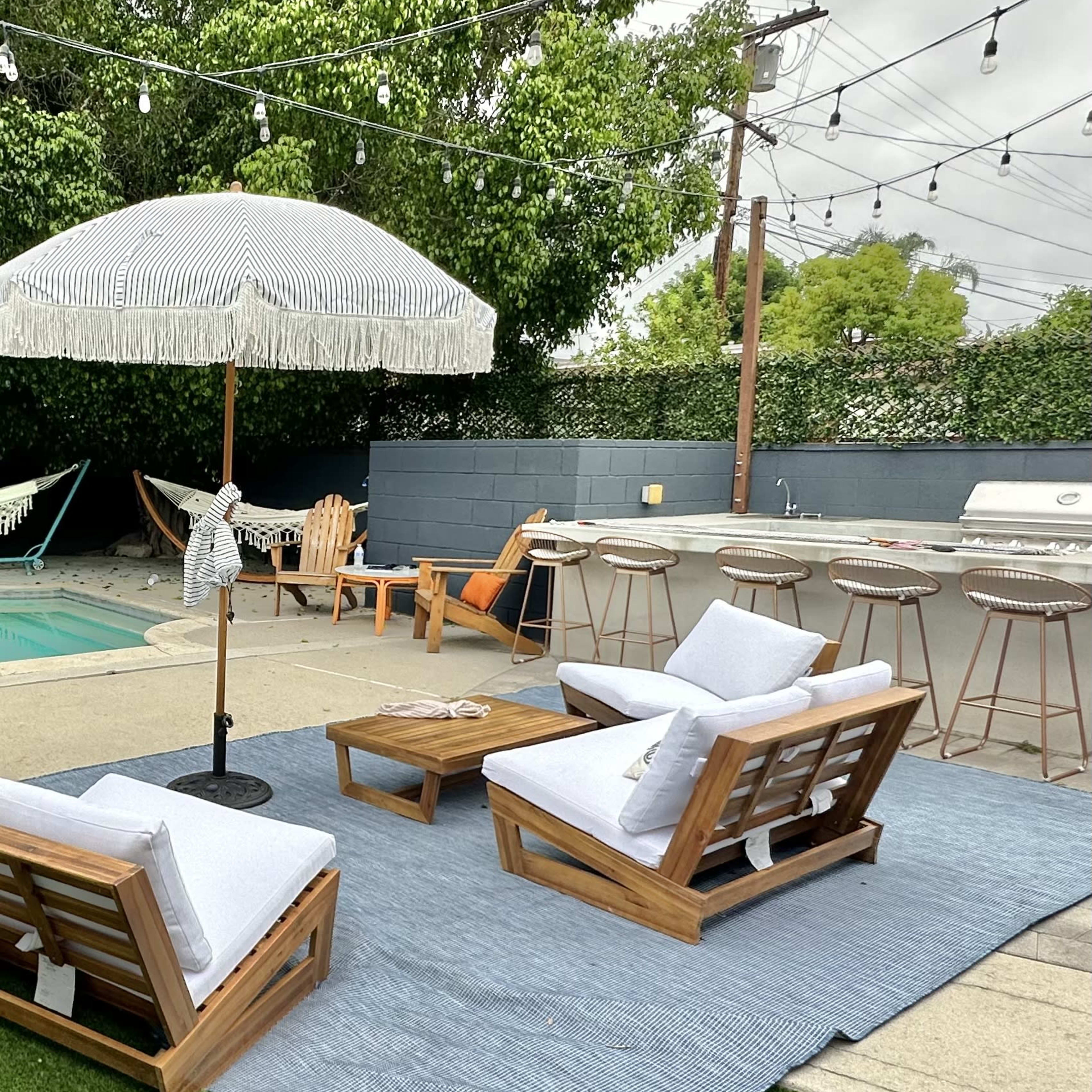 A backyard patio area with two wooden lounge chairs, a table on a blue rug, and an umbrella, surrounded by grass and various outdoor furniture.