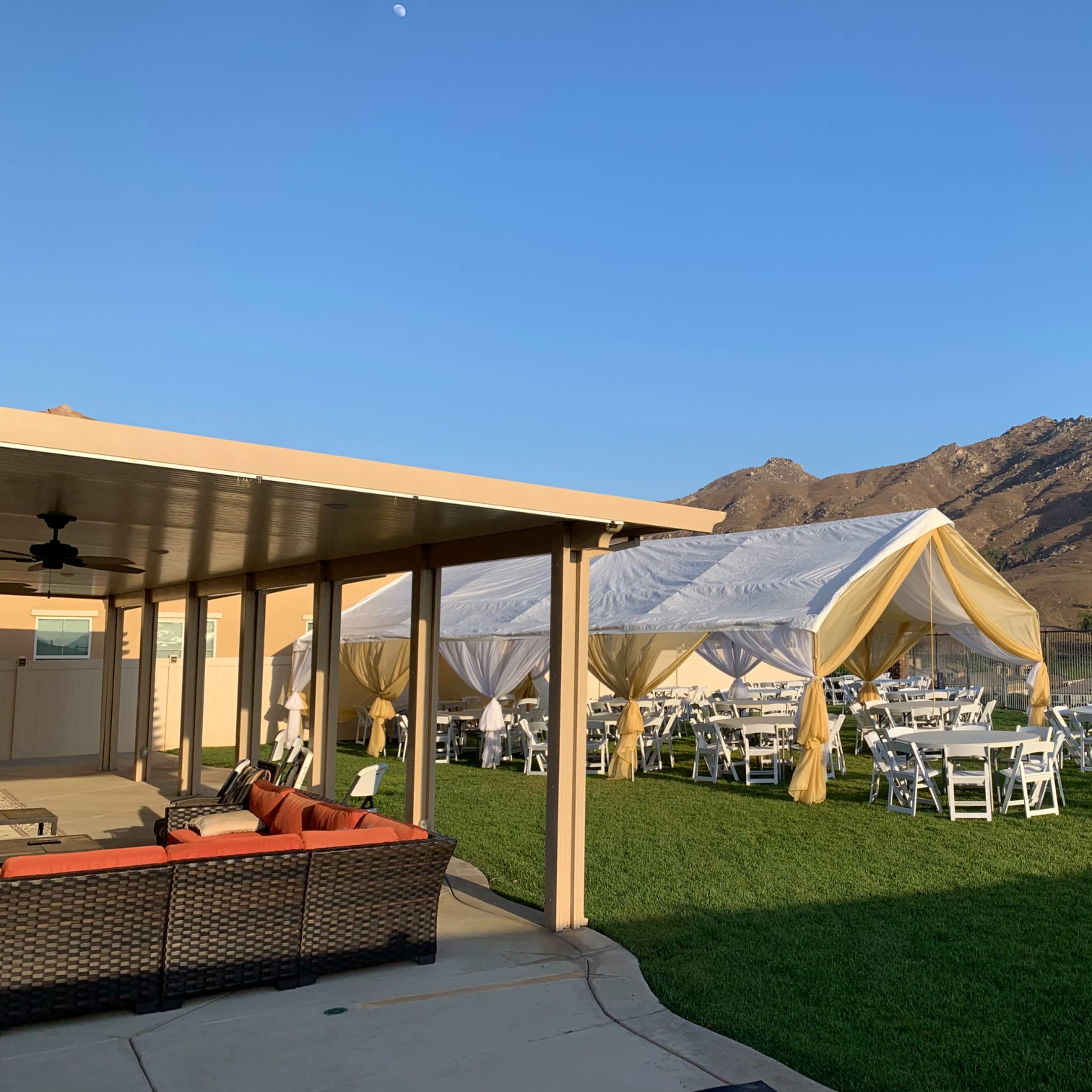 A white tent with yellow accents is set up in a grassy area next to a building, with tables and chairs arranged underneath, surrounded by mountains under a clear blue sky.