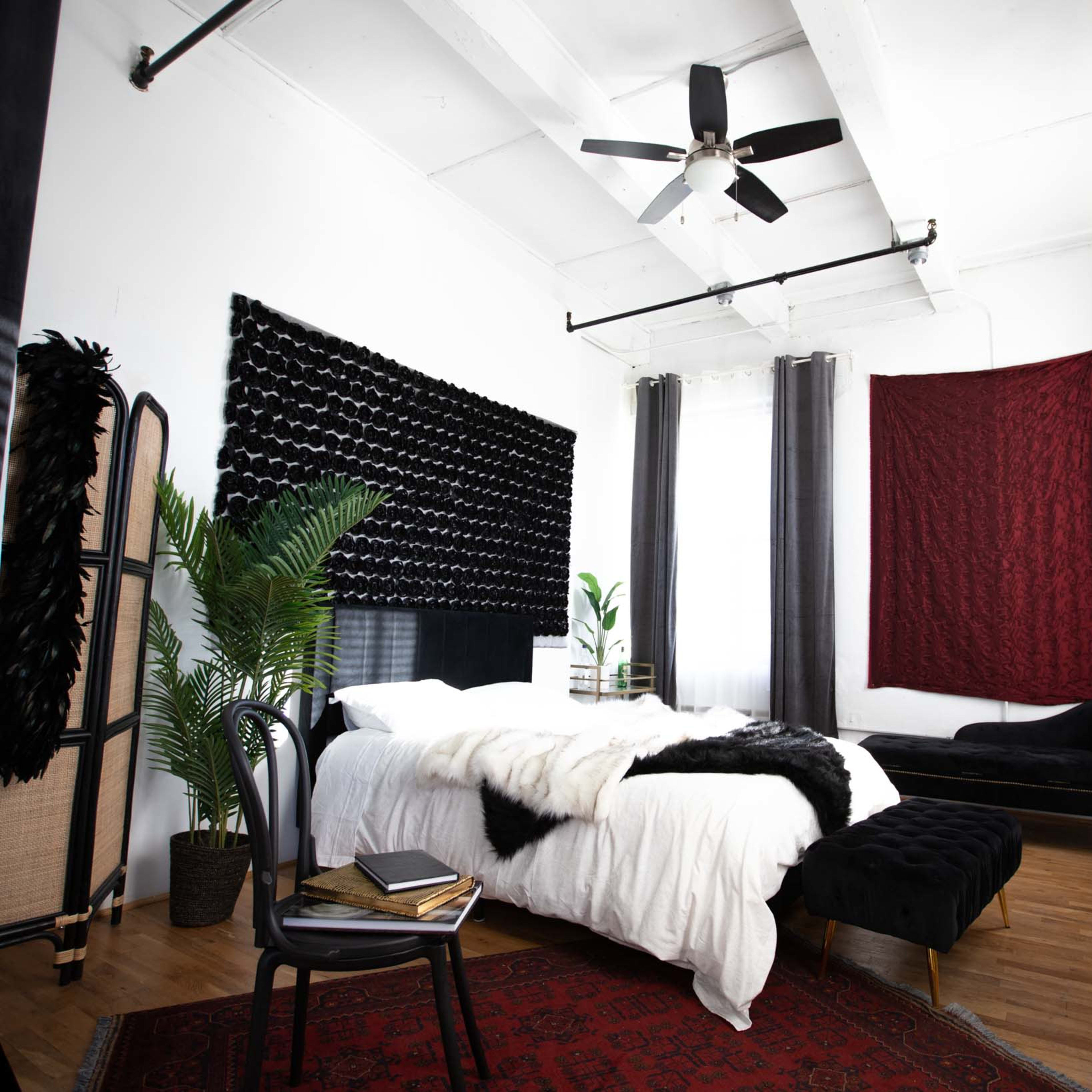The room features a bed with a black headboard, surrounded by potted plants and a vintage chaise lounge, all set against white walls and hardwood flooring.