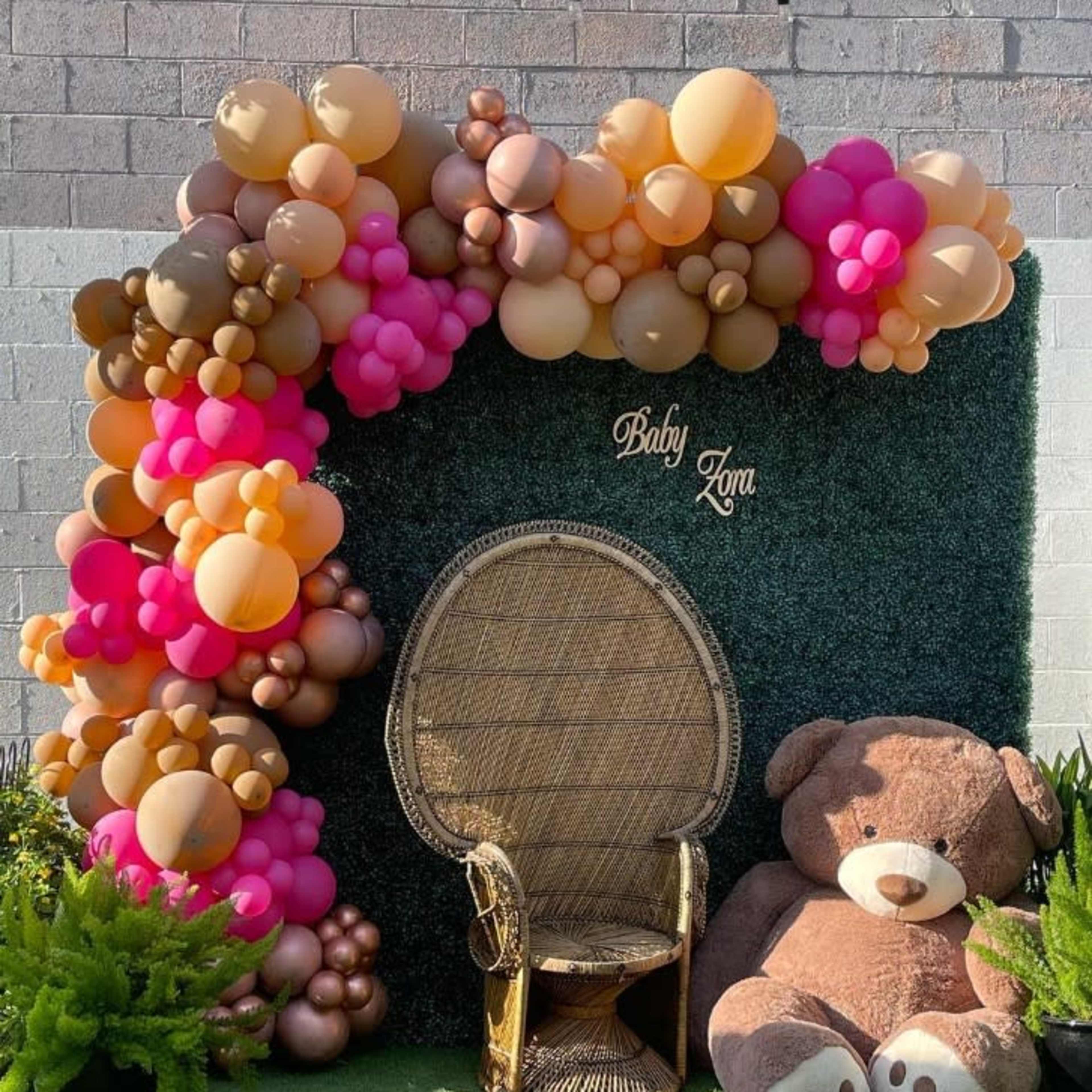 A decorative backdrop with an arch of colorful balloons and a plush teddy bear beside a gold chair.