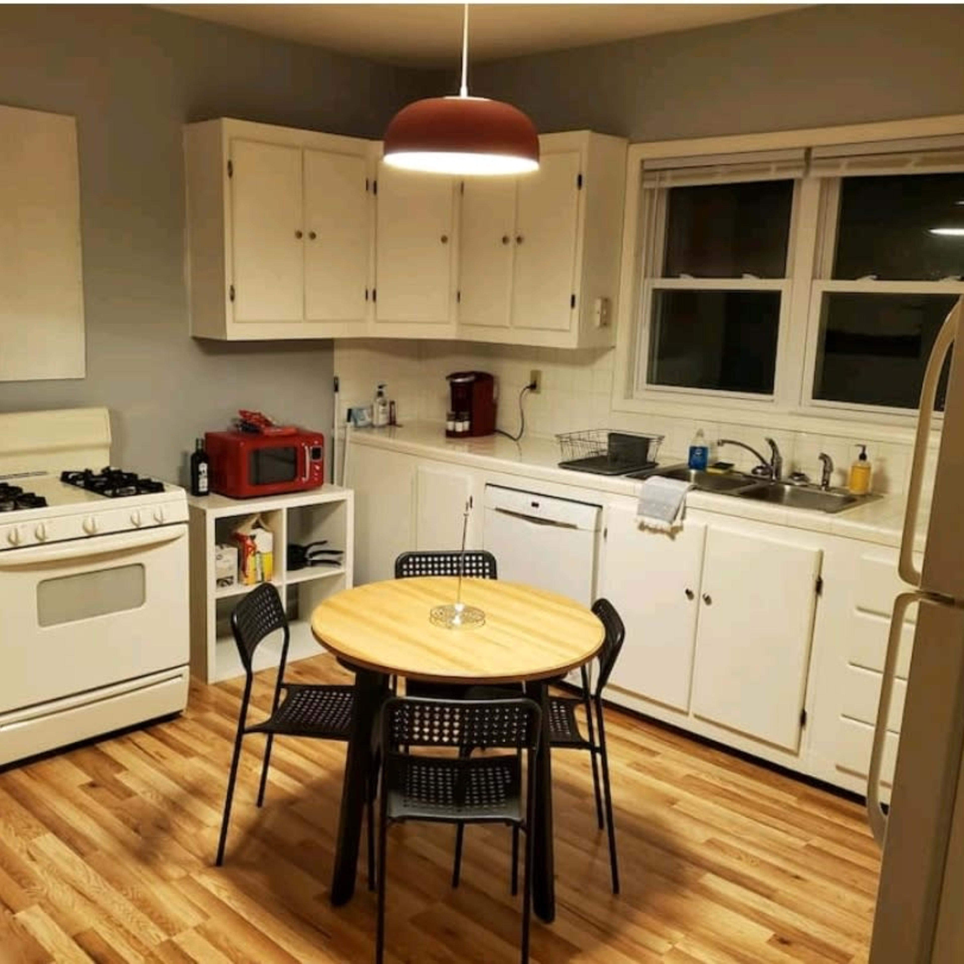 A kitchen with white cabinets, a round wooden table surrounded by four black chairs, a stove, and a sink beneath a window.