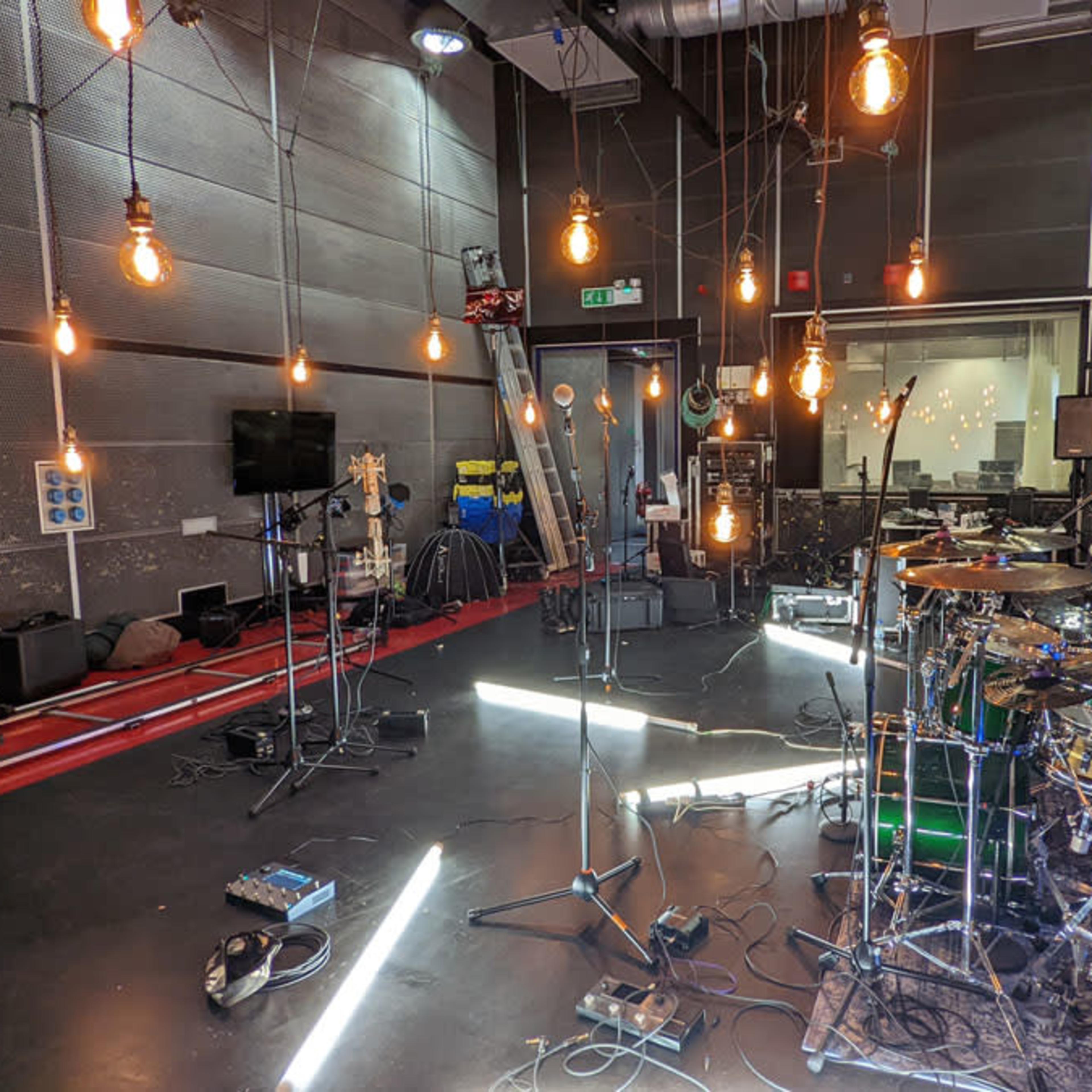 A music recording studio is illuminated by hanging light bulbs, featuring a drum set, microphones, and various equipment scattered around the floor.