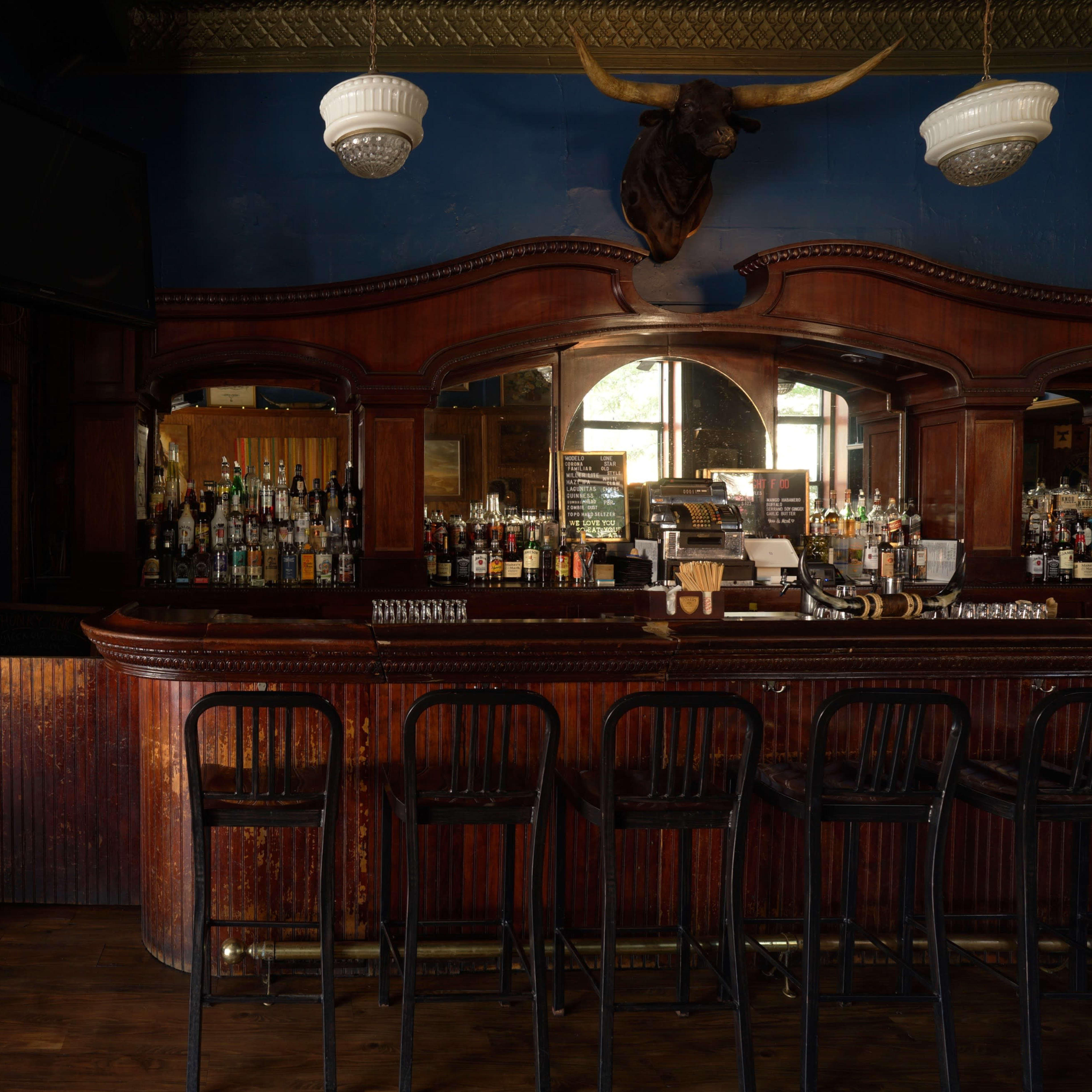 A dimly lit bar with a wooden counter, backlit shelves filled with bottles, and a mounted bull's head on the wall.
