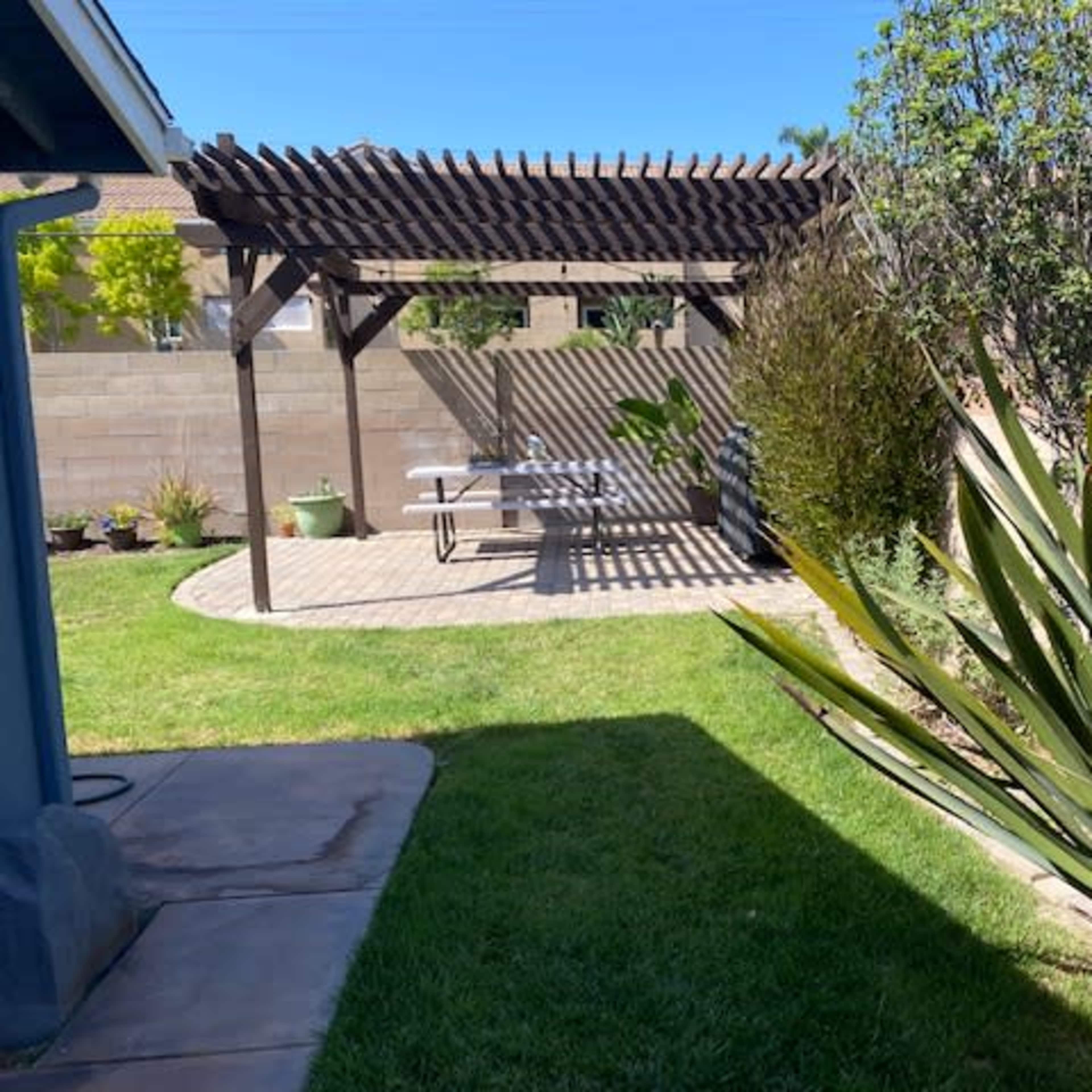 A backyard with a pergola, a stone patio with a picnic table, and landscaped greenery including large plants and grass.