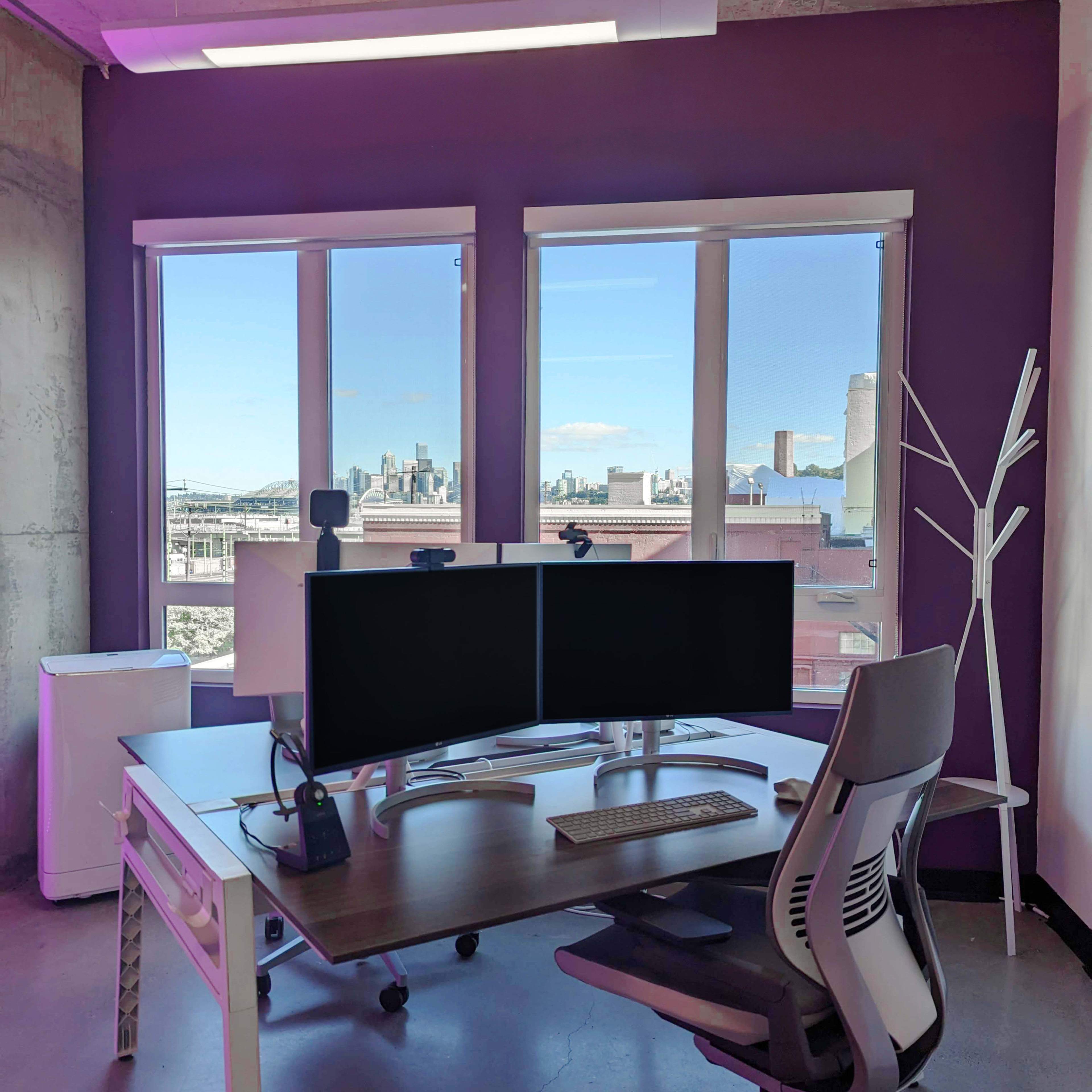 A modern office space features a desk with dual monitors, a keyboard, and a chair, set against a backdrop of large windows revealing a city skyline.
