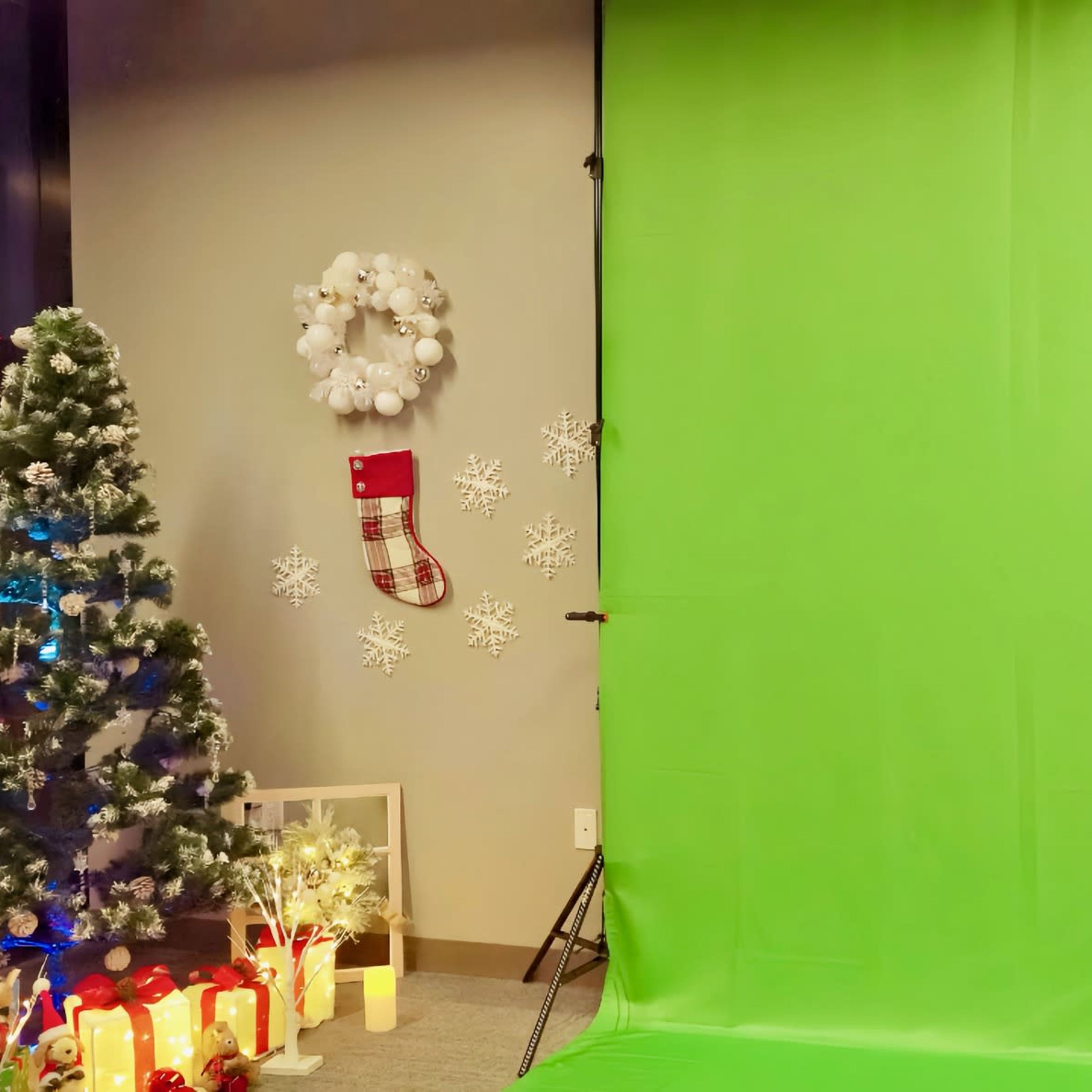 The scene features a green backdrop set up for photography, with a decorated Christmas tree, a wreath, a stocking, and snowflake decorations nearby.